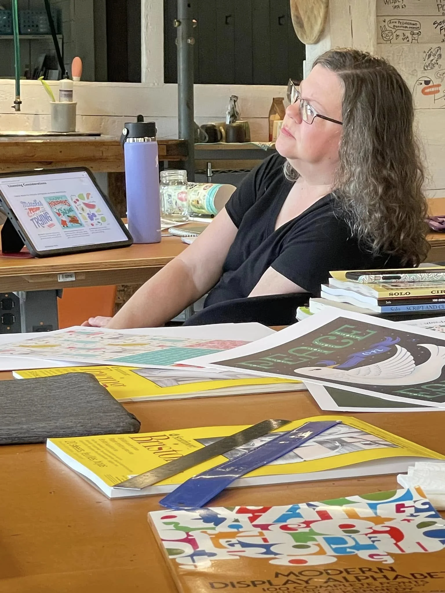 Artist listening to a demo at the studio table, with lettering reference books, printed alphabets and a tablet in front of her