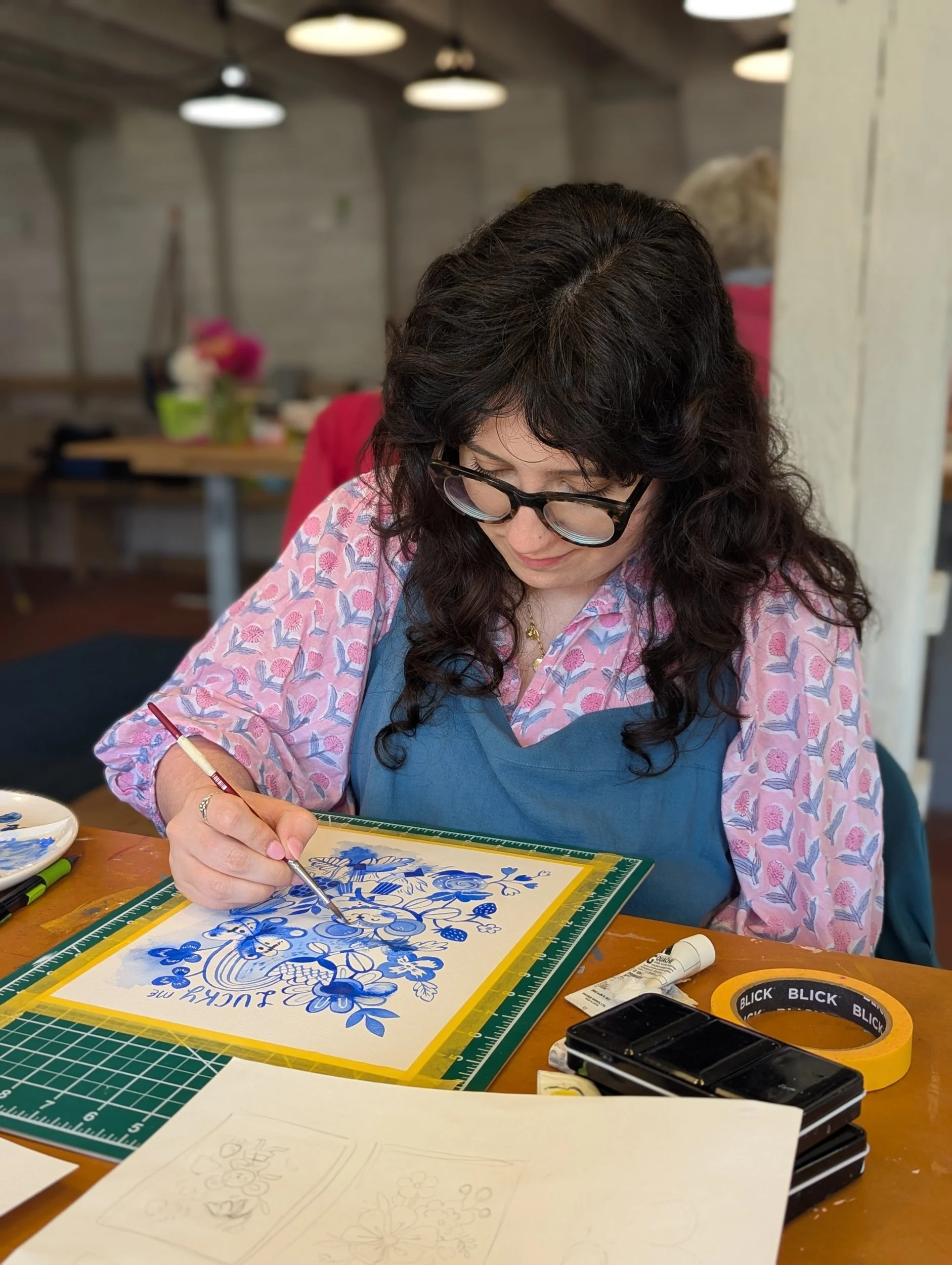 Artist painting a blue and white floral design on paper taped inside a frame at the barn studio table