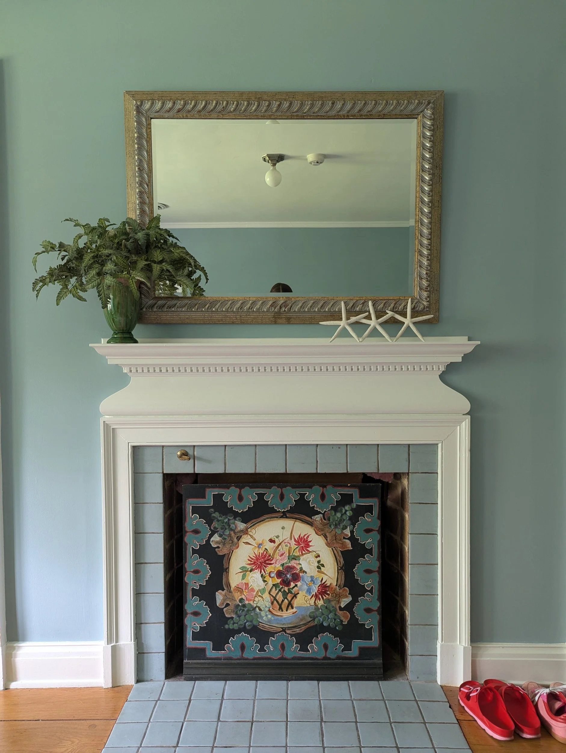 Fireplace in a sage green room with a painted floral fire screen, a gold framed mirror and a potted fern on the mantel