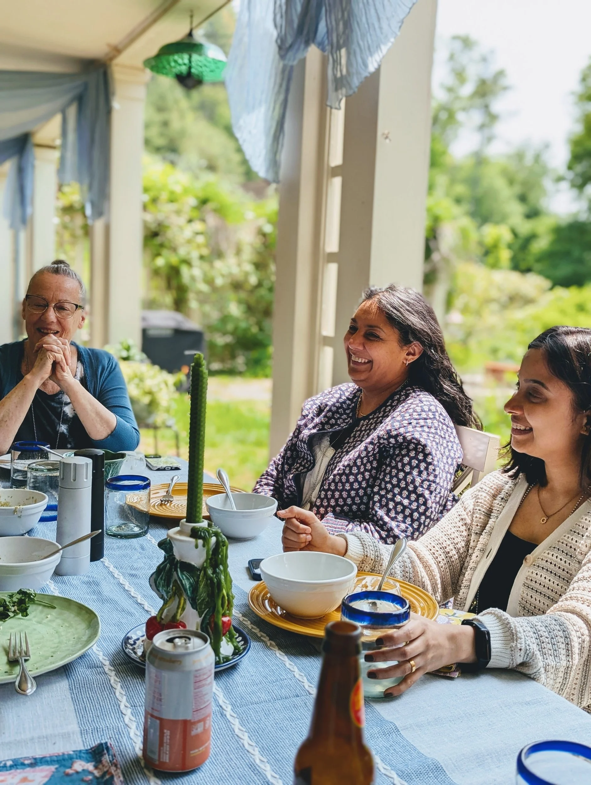 Three artists laughing together at a porch lunch table set with blue and white linens and a green candle