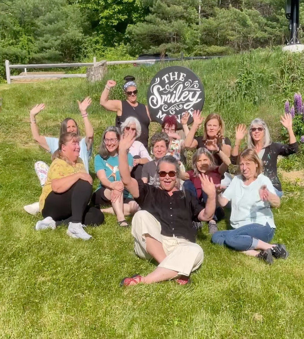 Group of smiling artists cheering with arms raised on the sunny hillside beside The Smiley Manse sign