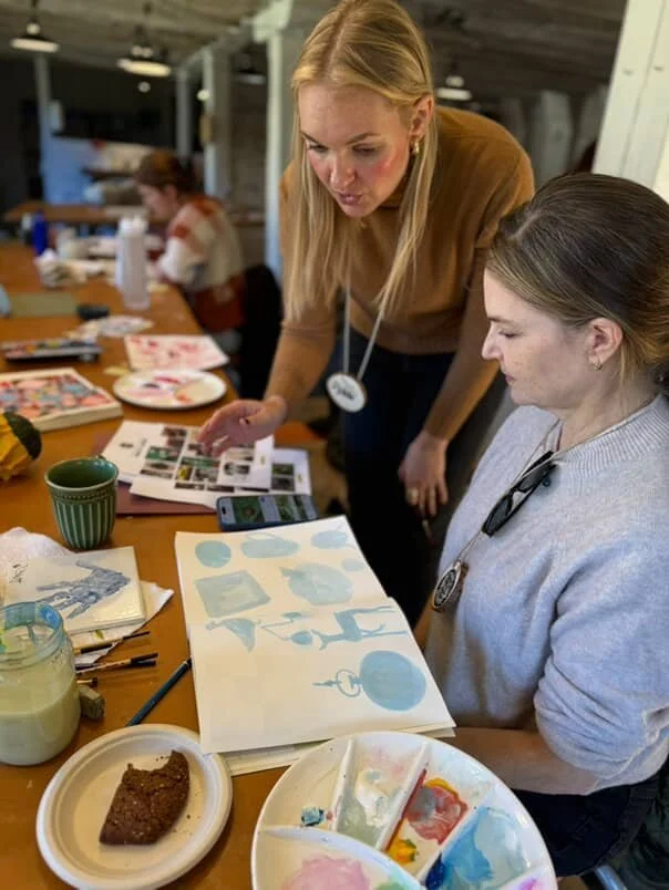 The instructor leans over a table to help an artist painting blue watercolor shapes in the barn studio