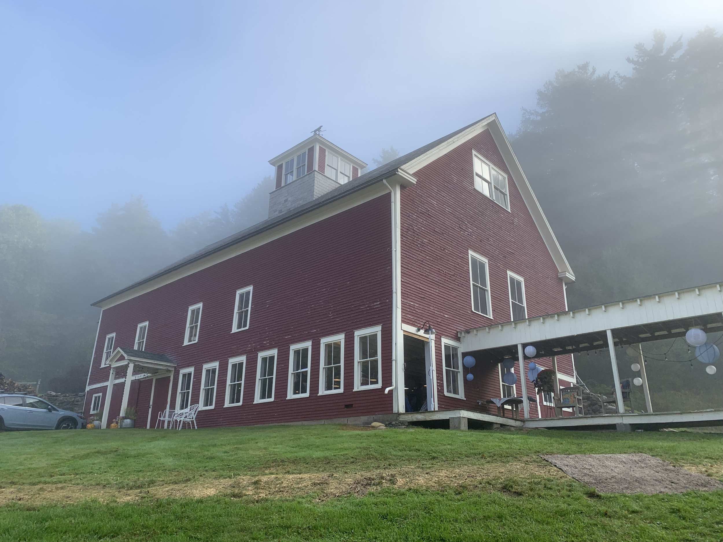 Red barn with a cupola and a covered porch hung with paper lanterns, softened by morning fog