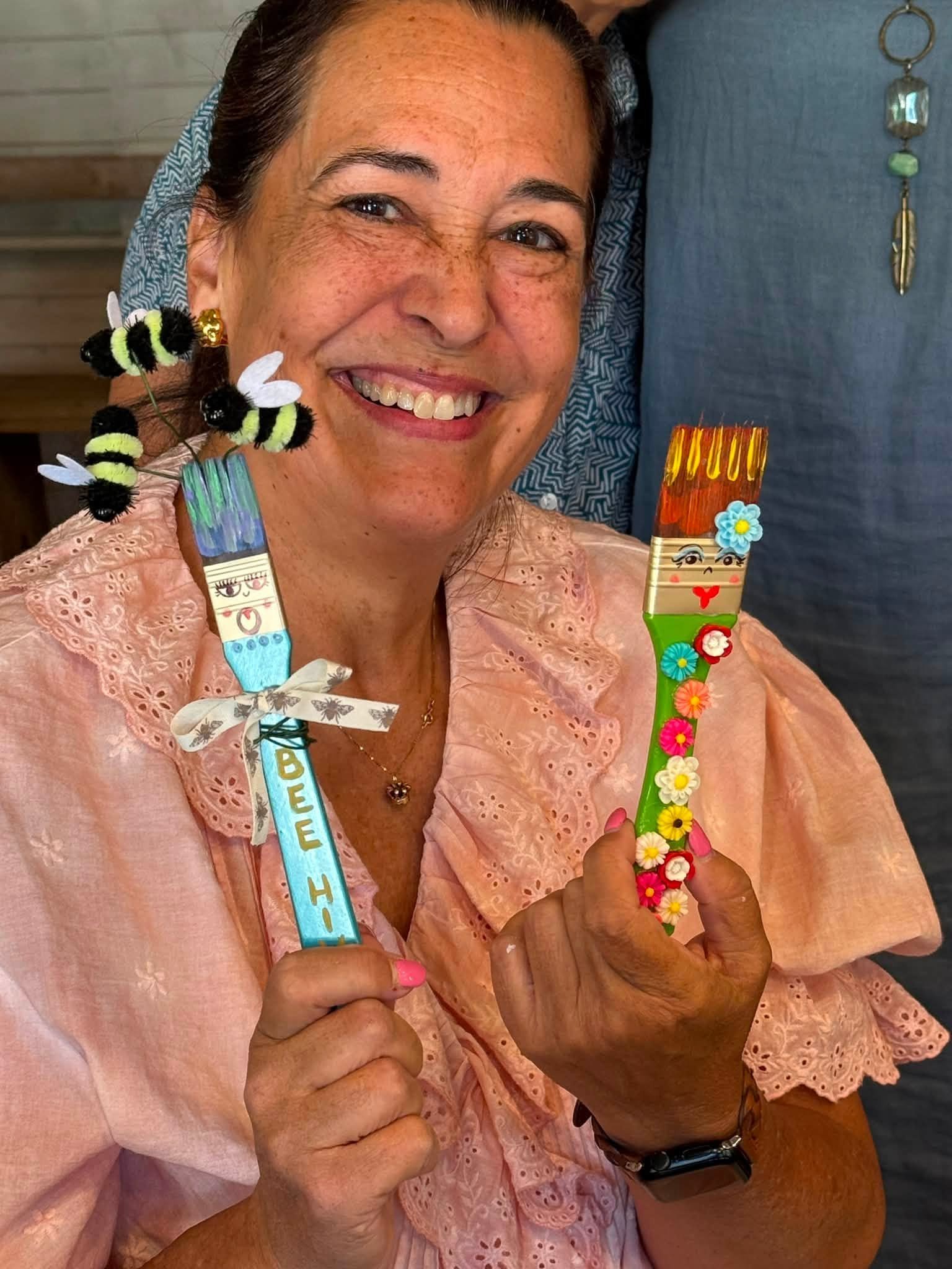 Smiling retreat artist holding two handmade decorated paintbrush sculptures with tiny bees and flowers