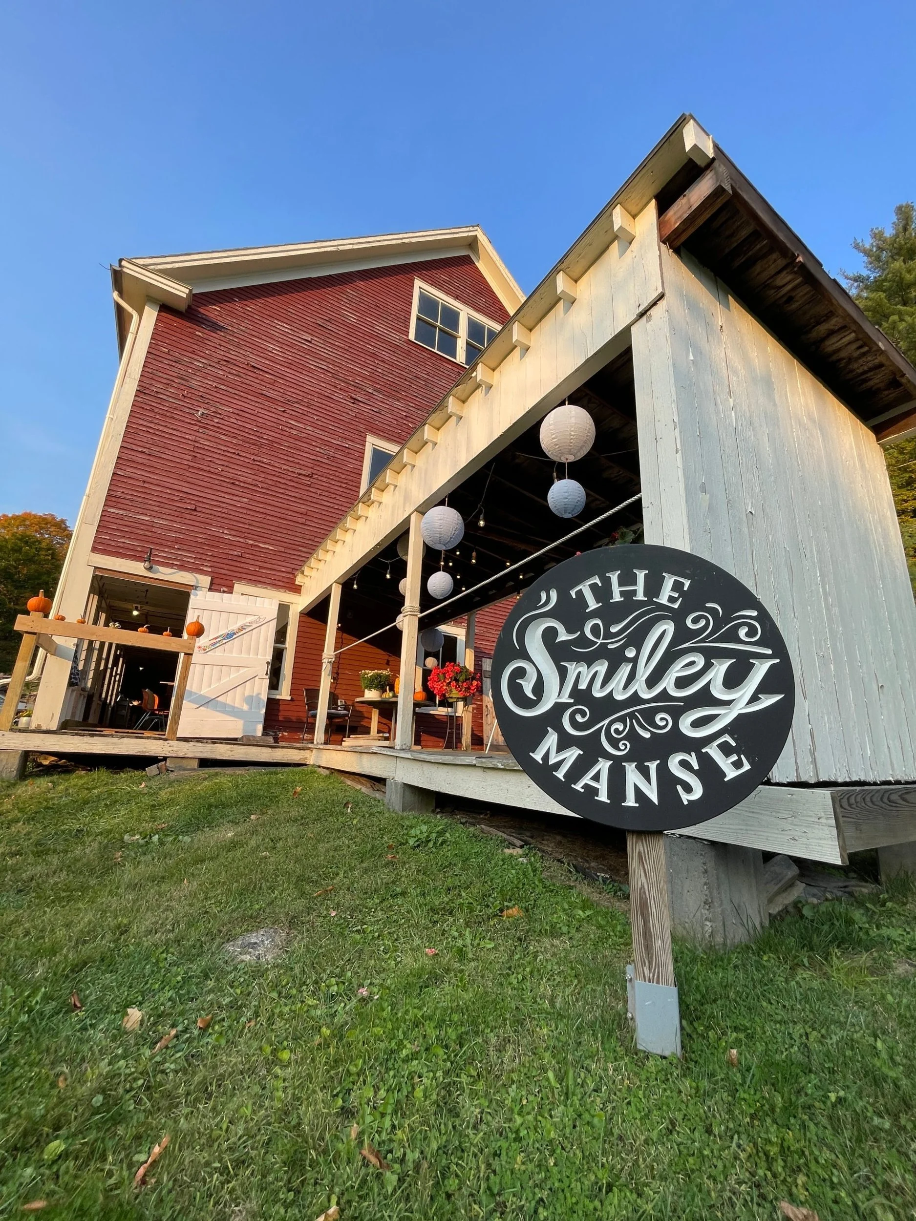 The round black and white The Smiley Manse sign on the lawn in front of the red barn with its porch hung with paper lanterns and pumpkins on the railing