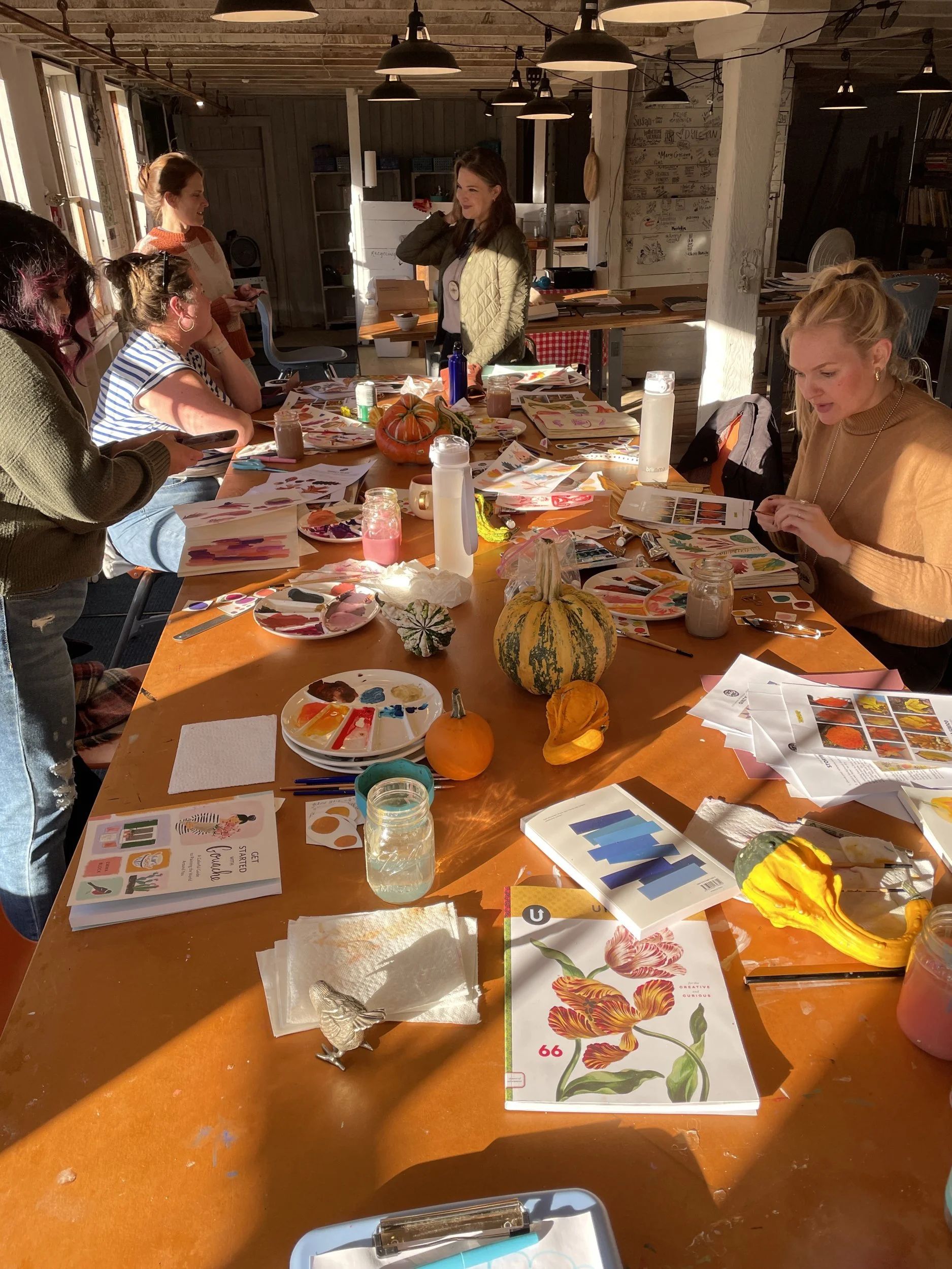 Long barn studio table covered with watercolor palettes, floral paintings, pumpkins and gourds