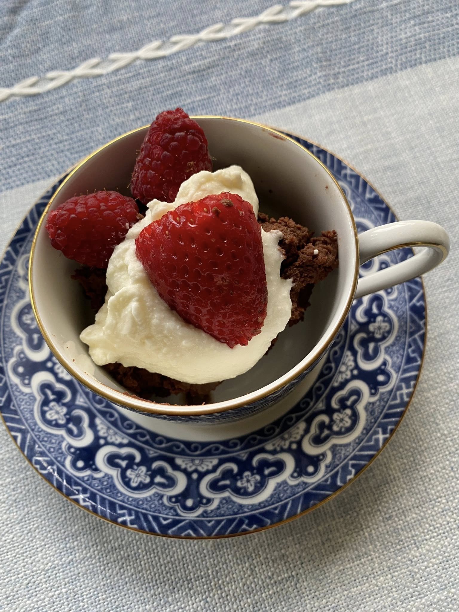 Chocolate cake topped with whipped cream, a strawberry and raspberries served in a blue and white teacup