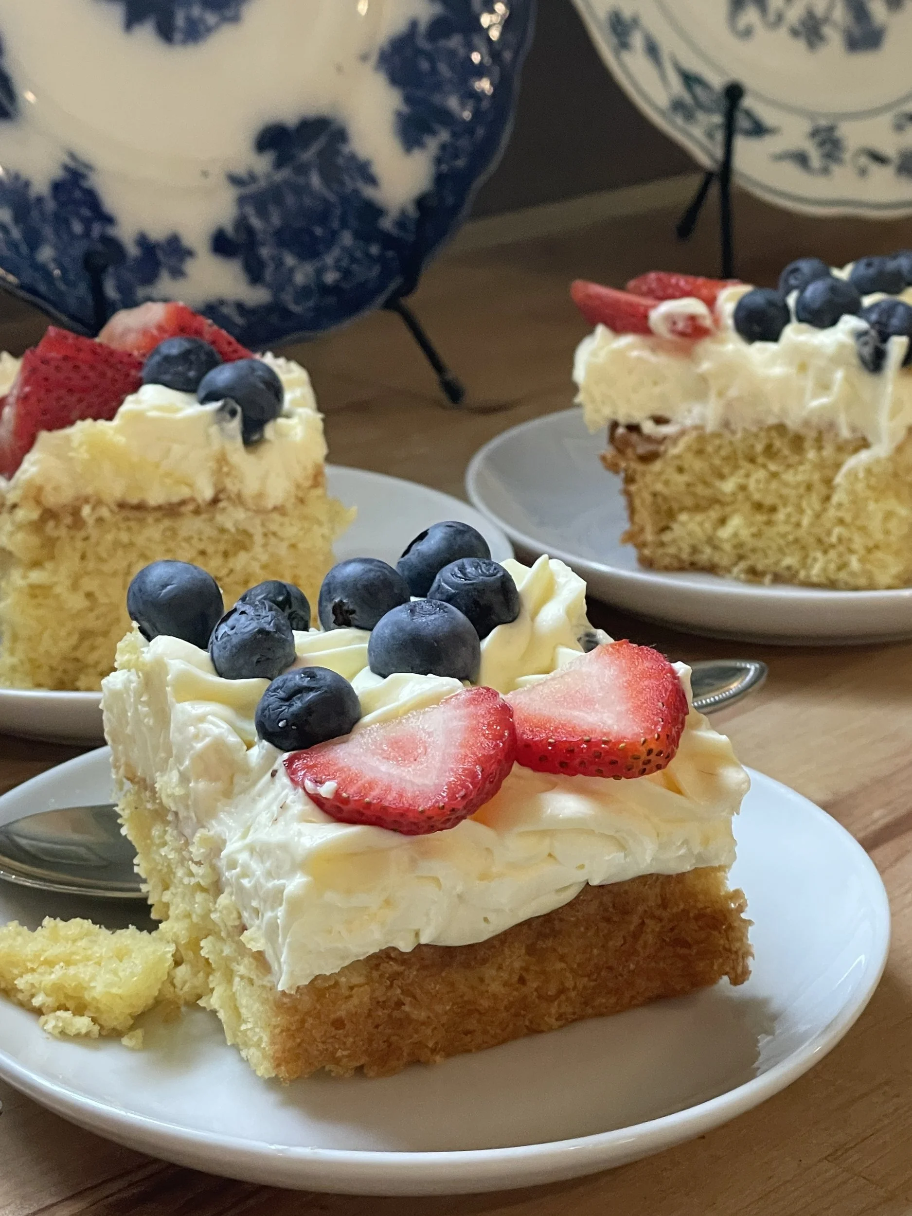Slices of berry-topped cream cake on white plates with blue and white china in the background