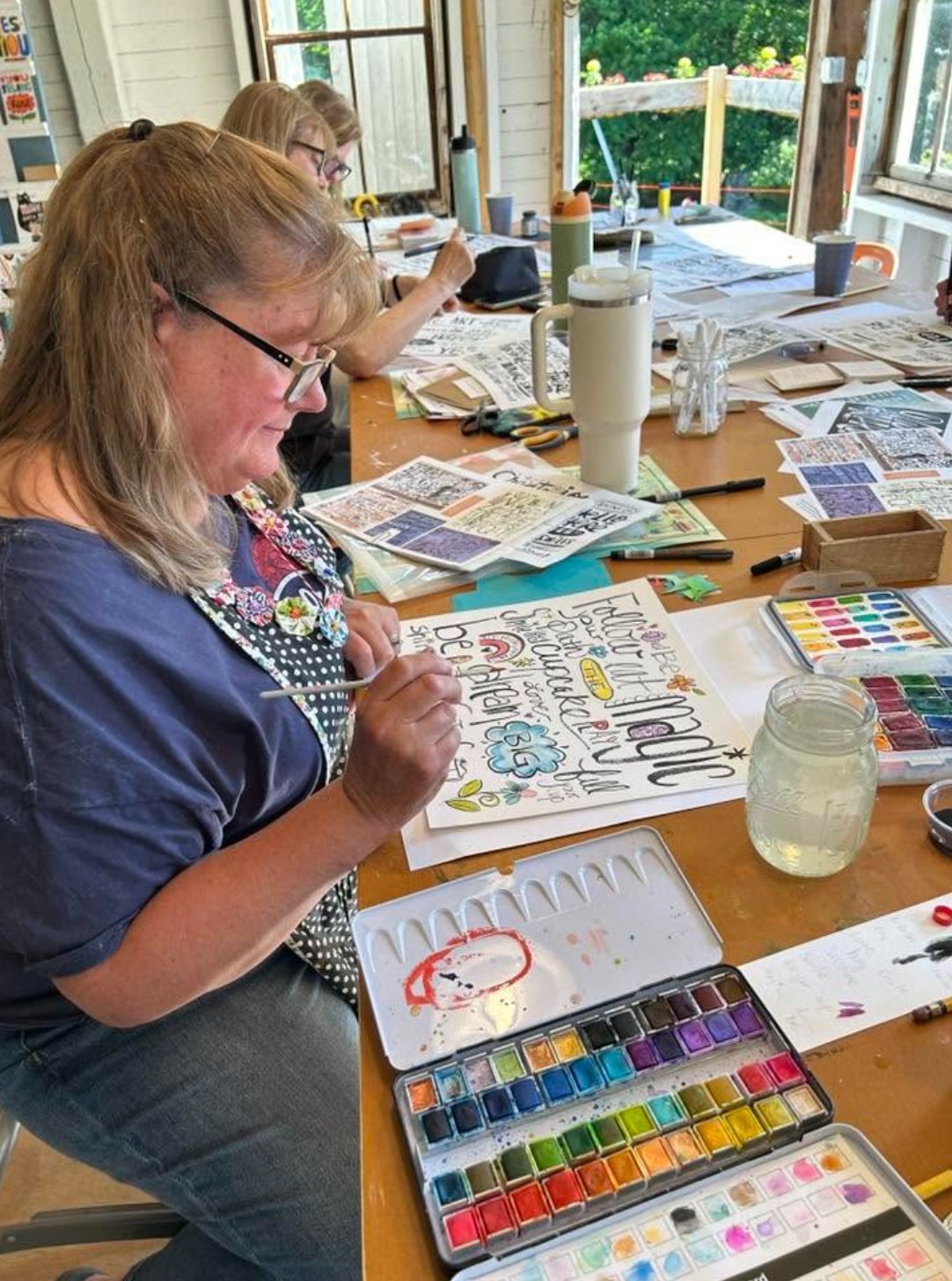 Artist painting hand lettering with a watercolor palette and jar of water at a studio table covered in reference sheets.