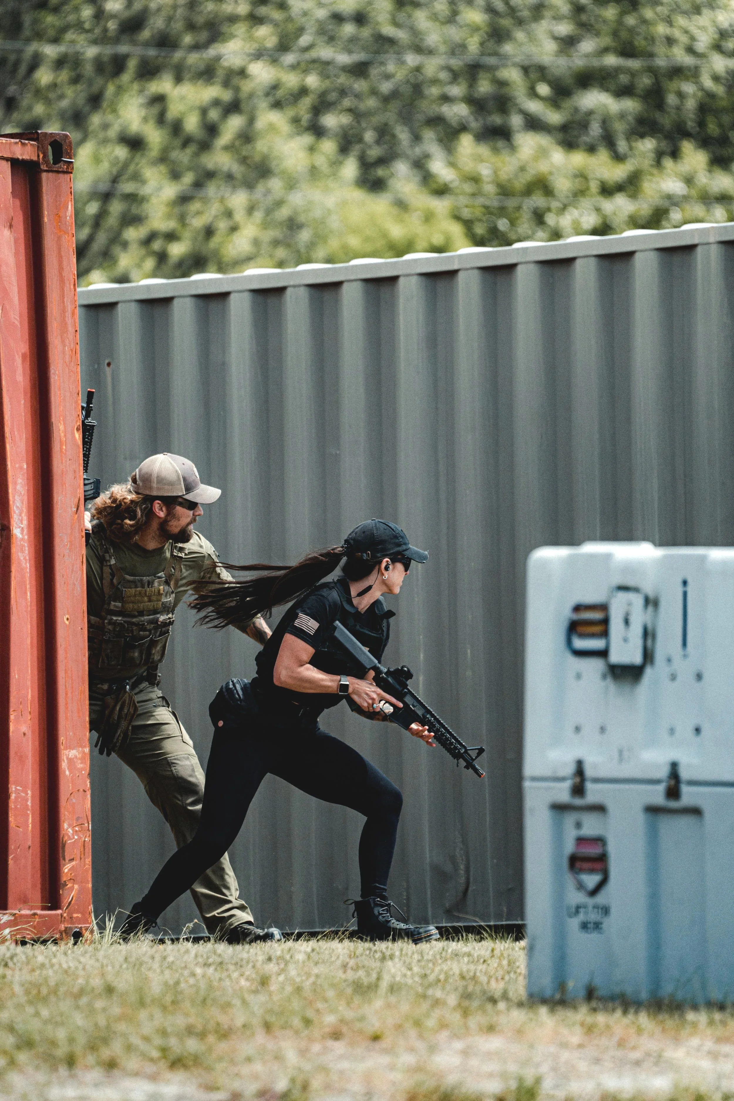 Two people, a woman and a man, participating in a tactical training or simulated combat scenario outdoors. The woman is crouched, holding a rifle, and the man is positioned behind her, seemingly guiding or protecting her. The scene is set near a large gray fence and a red structure, with a grassy area and trees in the background.
