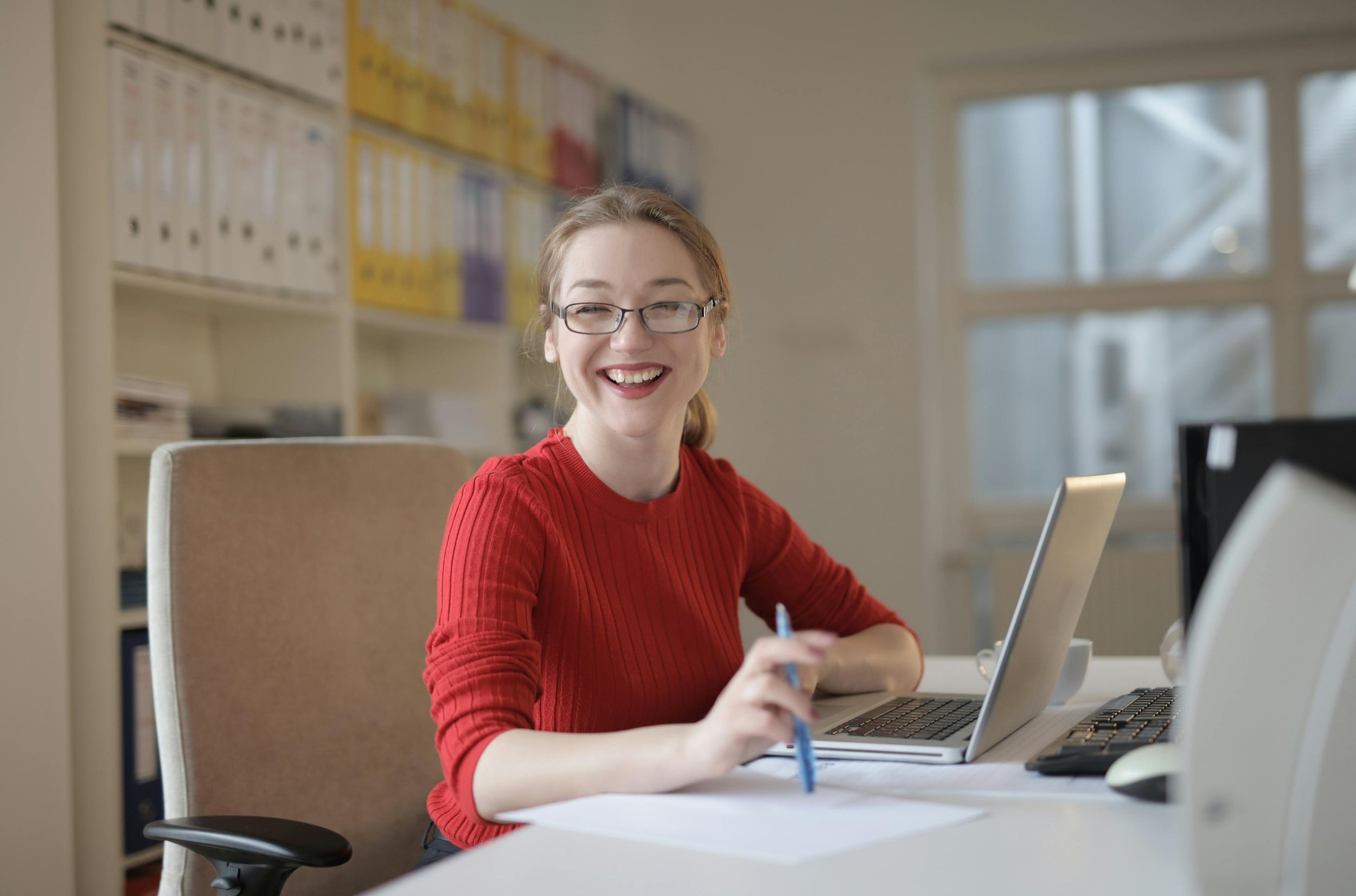 A young woman with glasses and red sweater sitting at an office desk, smiling, with a laptop and pencil in hand, in a bright office with bookshelves and windows in the background.