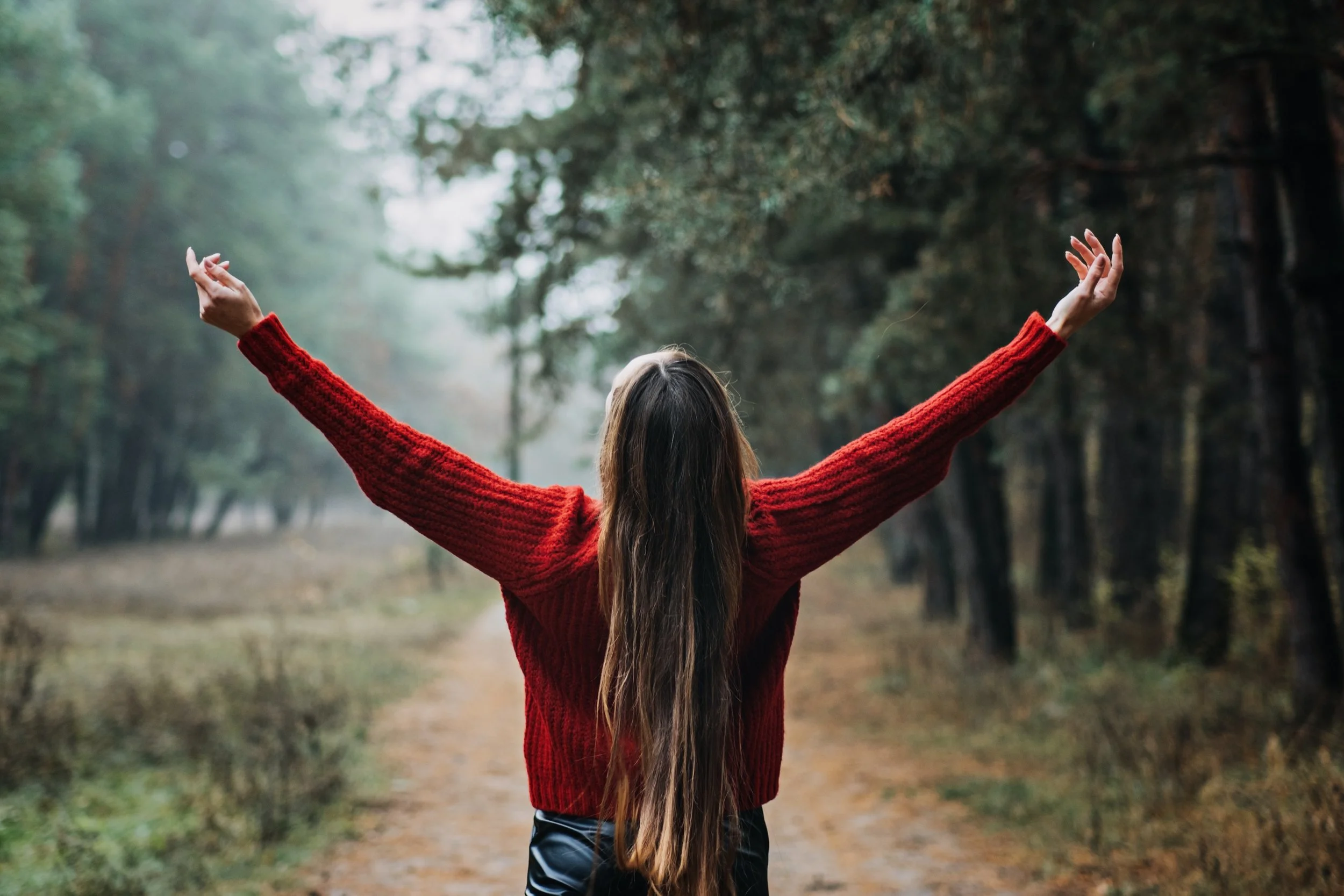 A person with long hair, wearing a red sweater, has their back to the camera with arms outstretched, standing on a dirt path in a forest surrounded by trees and greenery.