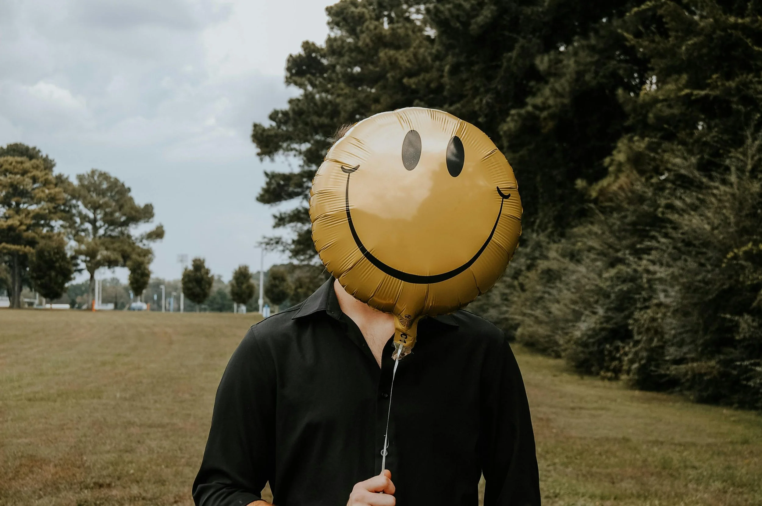 Person holding a yellow smiley face balloon in an outdoor park area with trees and cloudy sky in the background.