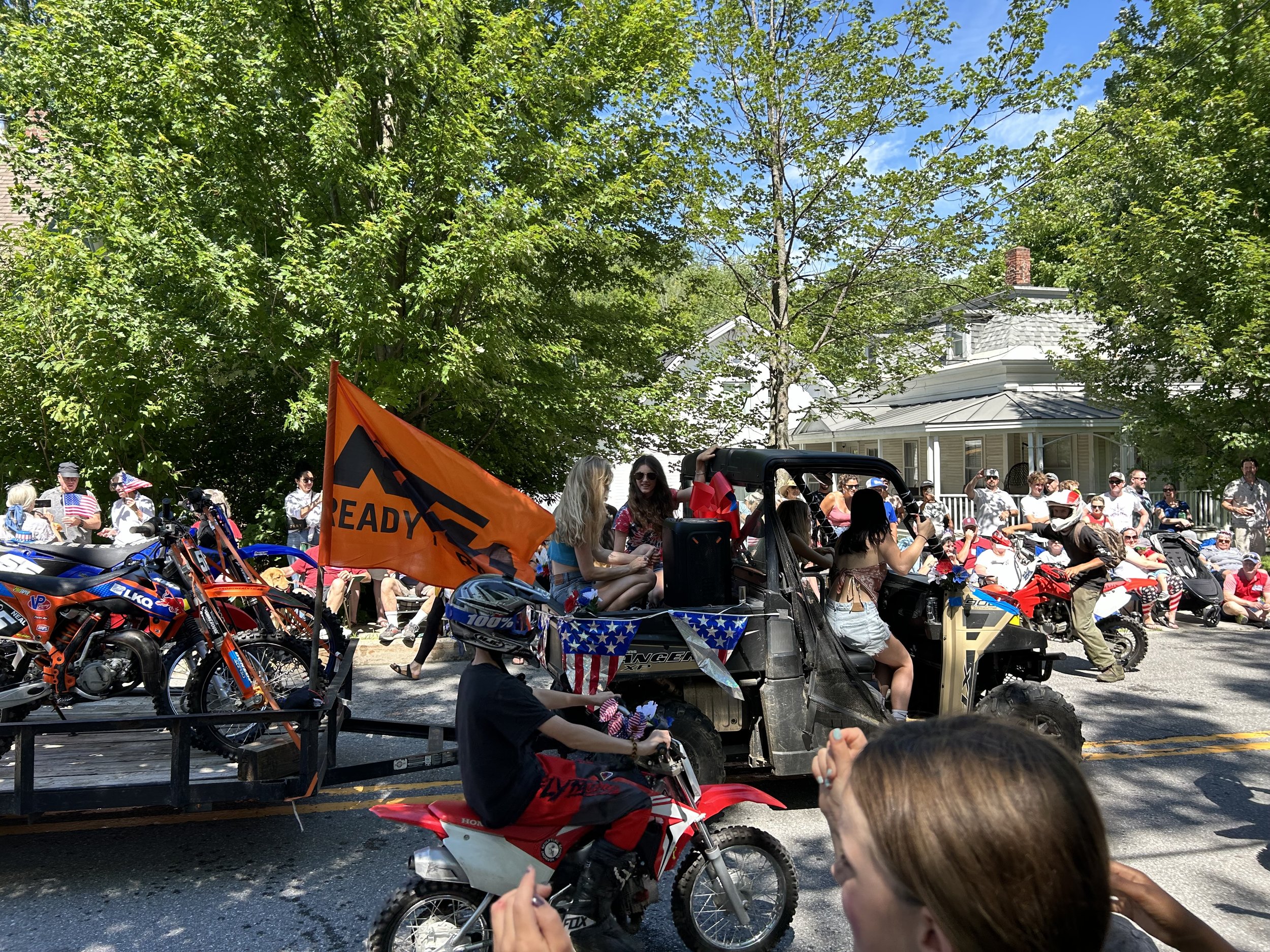 Group of people at an outdoor parade or gathering, some sitting on riding lawn mowers decorated with patriotic flags, and others standing or seated nearby. There are trees and houses in the background, and a person holding a flag that says 'READY'.