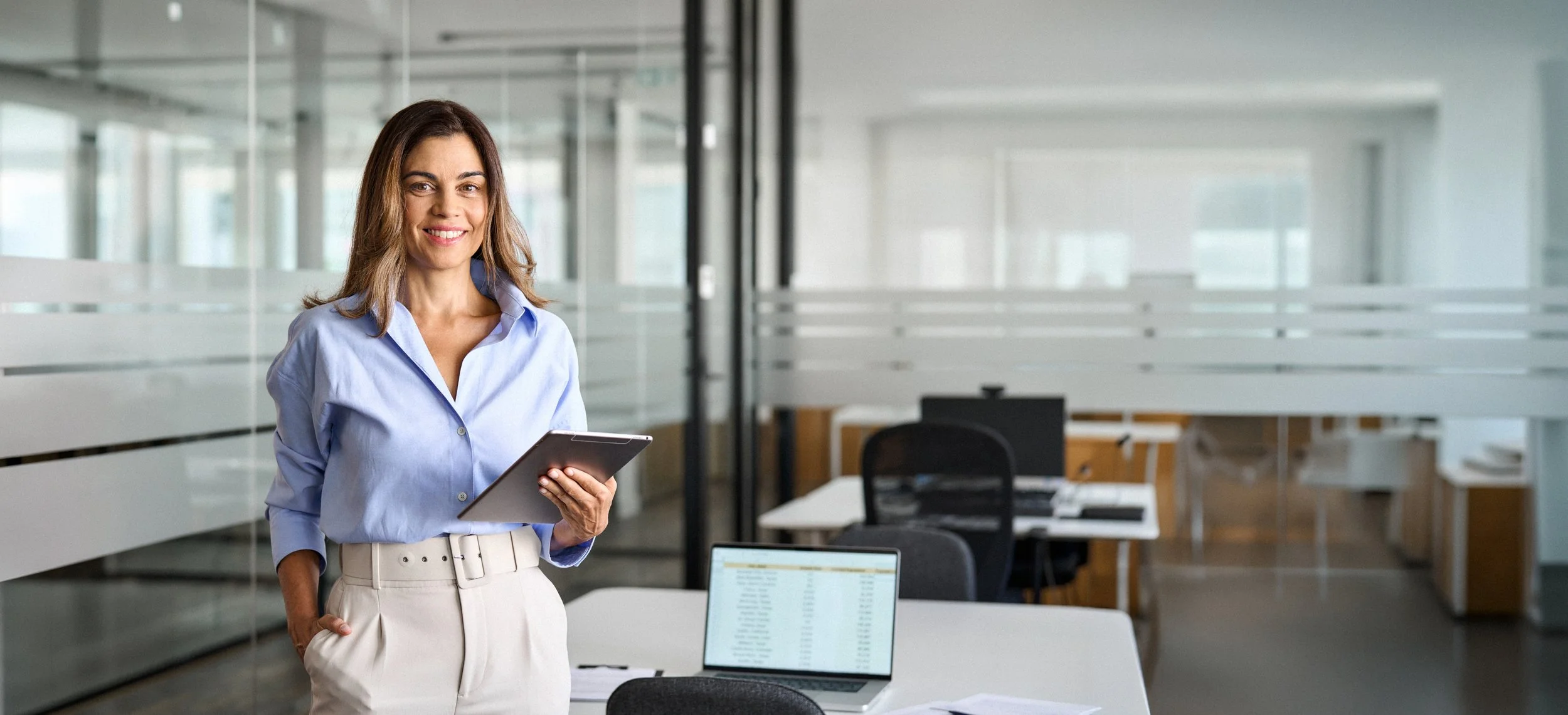 A woman standing in an office holding a tablet, with a laptop open on a desk in front of her.
