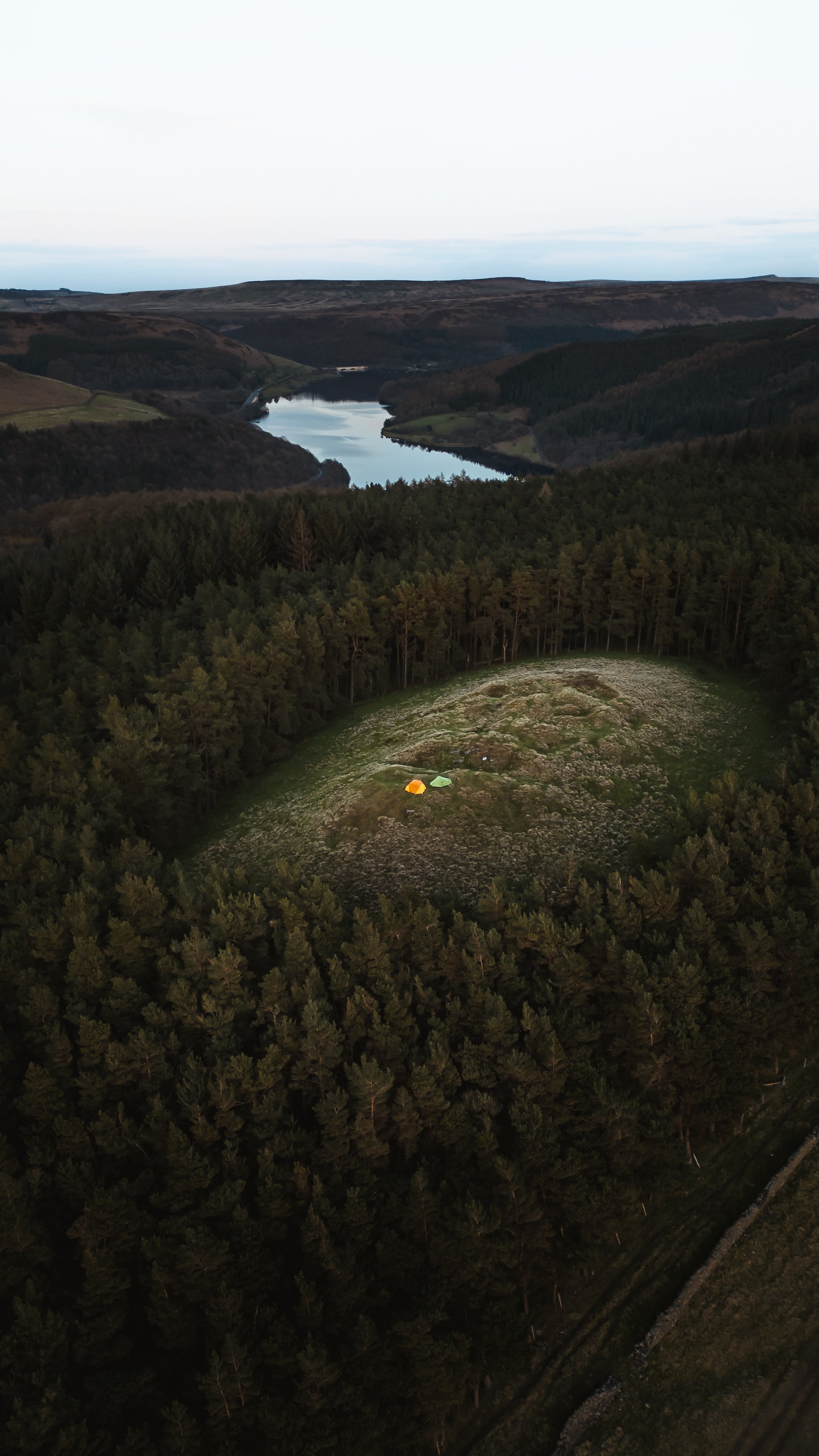 Aerial view of a forest clearing with two tents, surrounded by dense trees and overlooking a distant river.