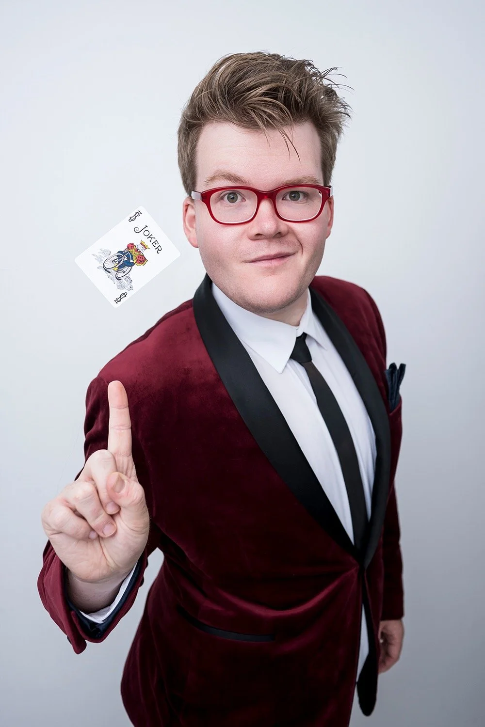 Australian Champion Magician Josh Staley in a red velvet tuxedo balancing a Joker playing card on his fingertip.