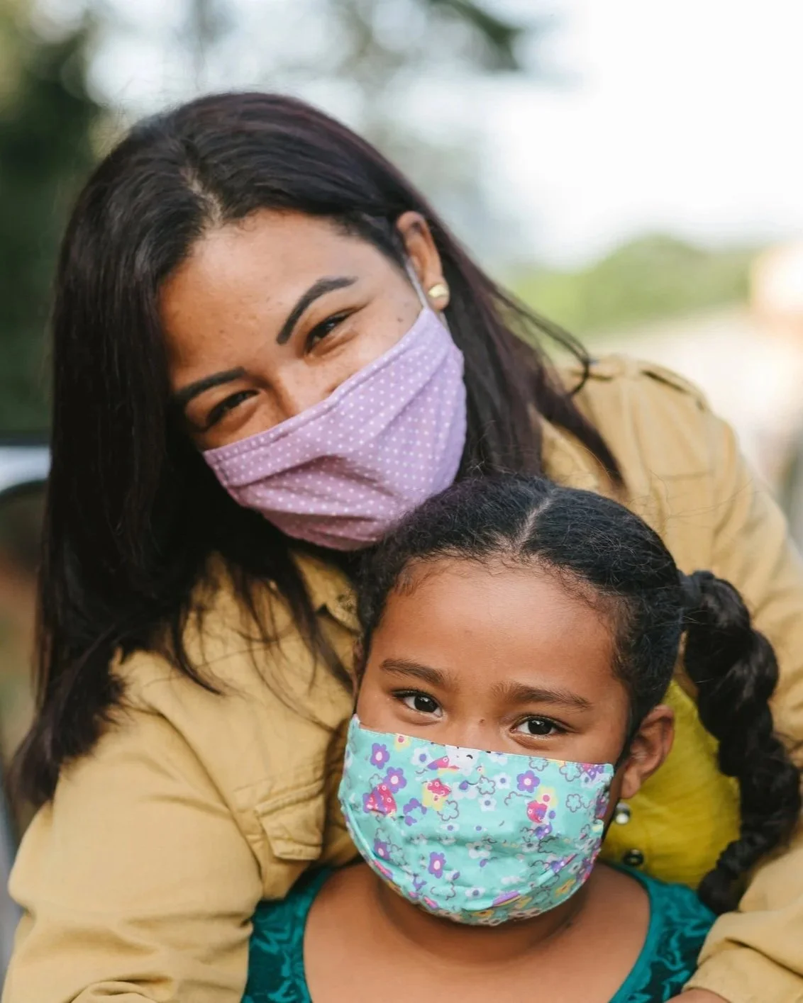 Photograph of a brown-skinned woman with long dark hair hugging a young child with braids. They are both wearing masks.