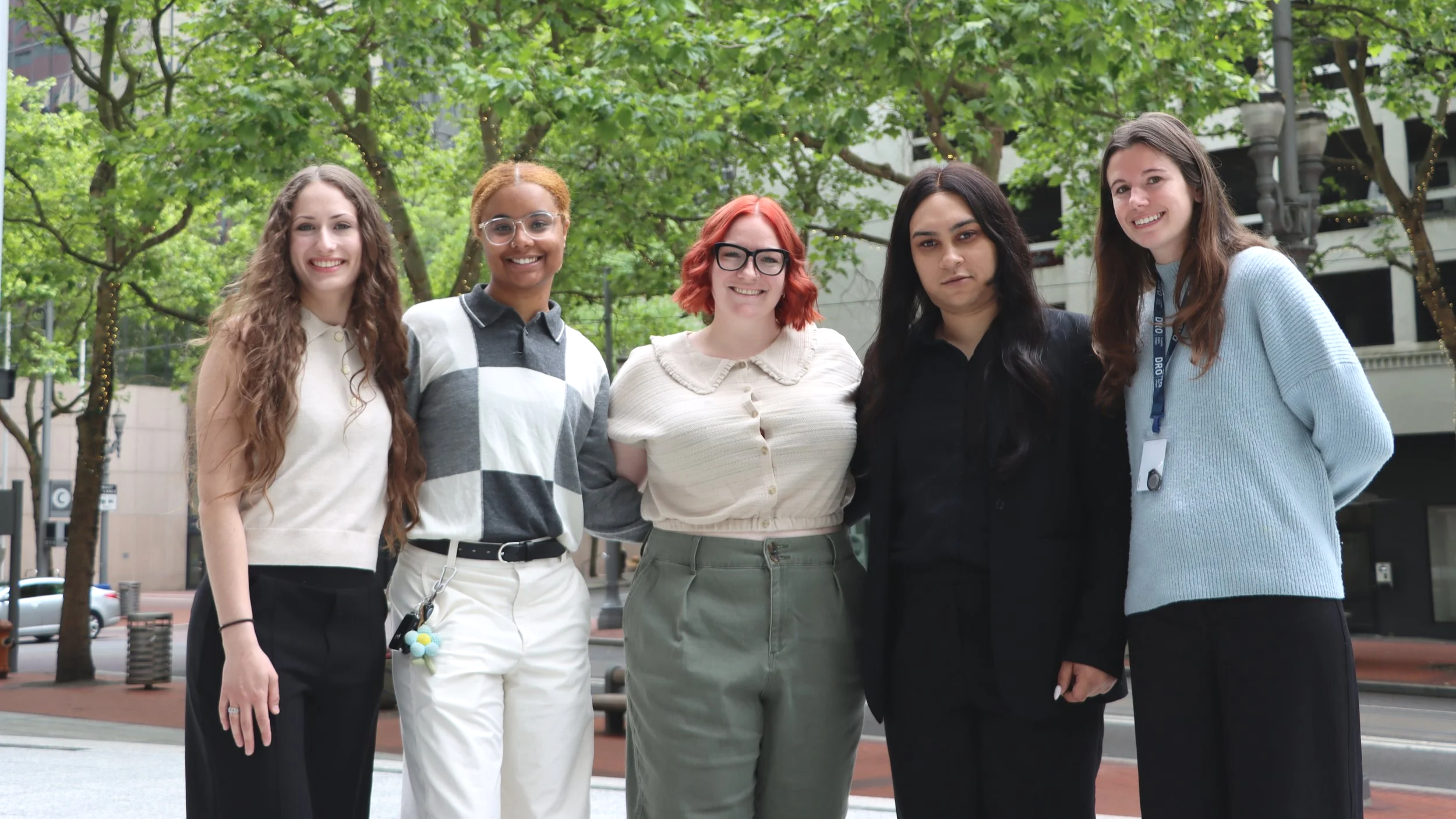 Five young law clerks standing outside in downtown Portland, Oregon, with buildings behind them. They are smiling with their arms locked together.