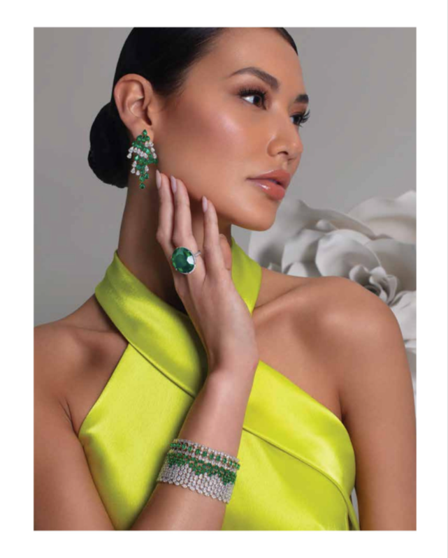 A woman with dark hair styled in a low bun wearing a bright yellow satin dress, large green gemstone ring, matching green and white earrings, and a sparkly bracelet. She touches her face with her right hand, with a neutral expression, against a gray 