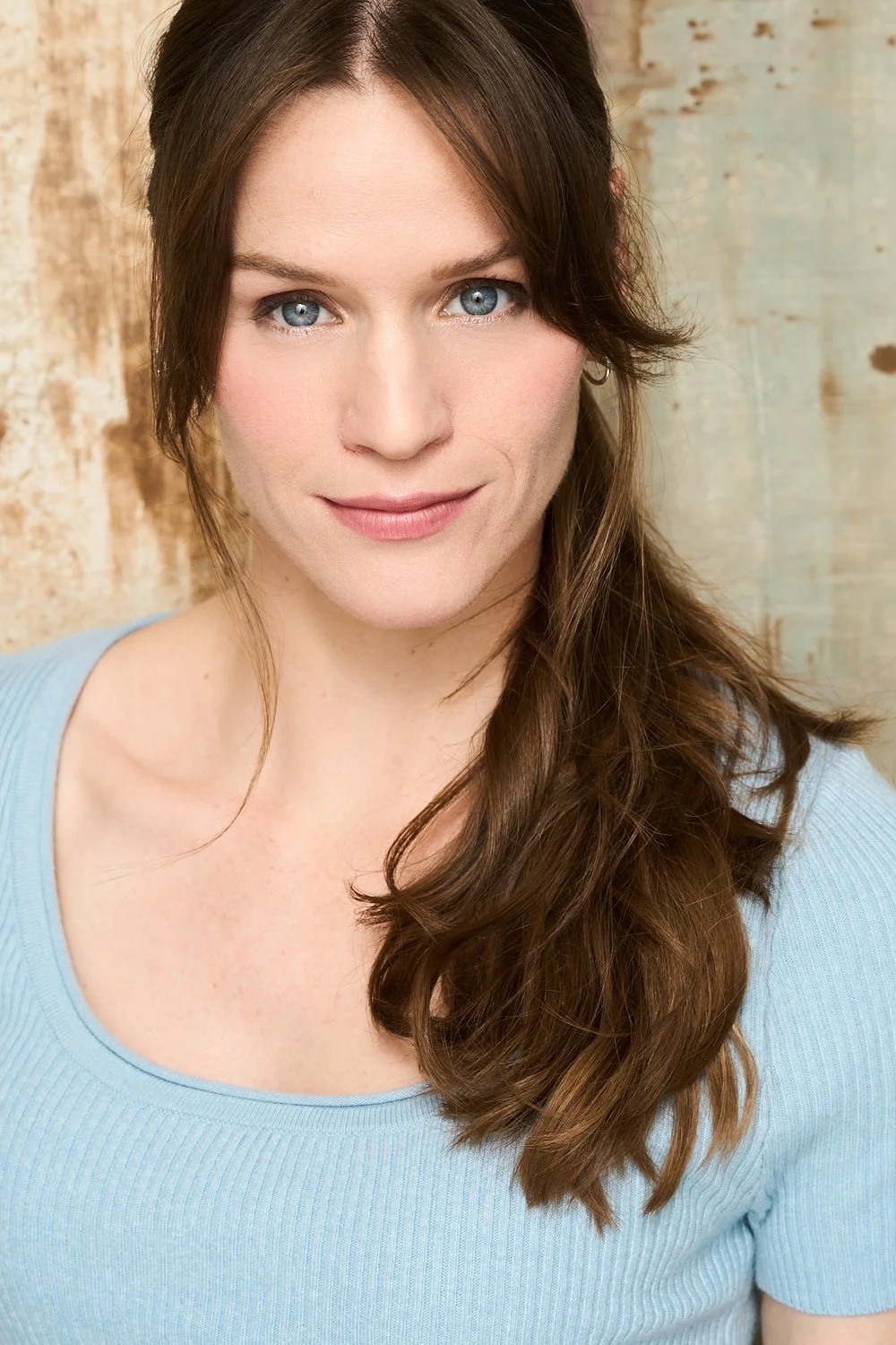 A woman with long brown hair and blue eyes smiling, wearing a light blue top, with a textured beige and light blue background.