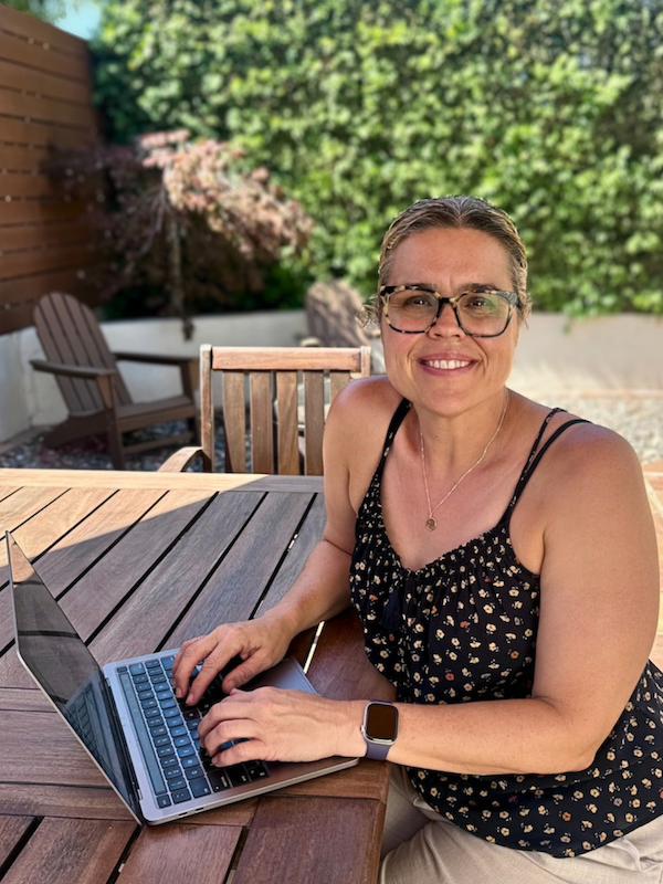 Woman smiling in a garden setting, wearing a black top, colorful pants, and a black cardigan.