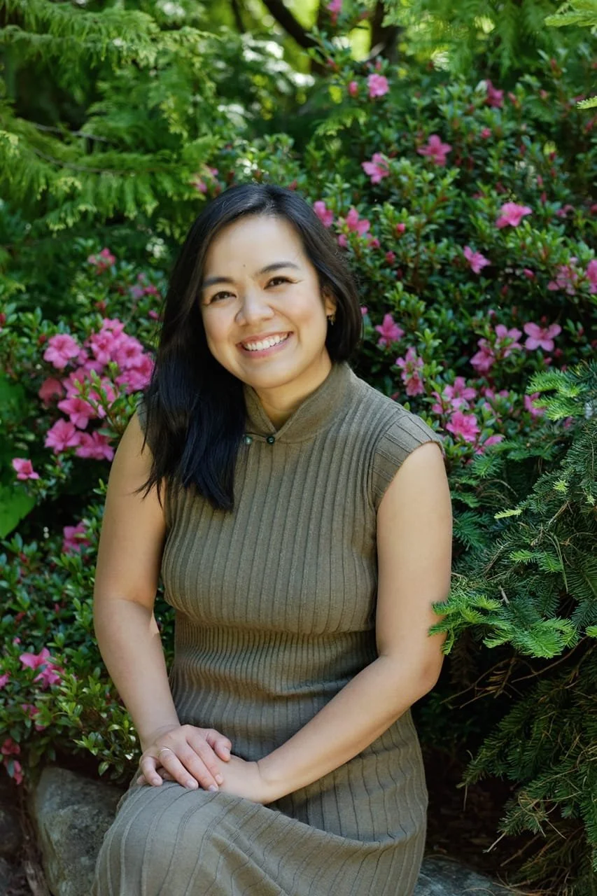 A young woman with black hair, smiling, sitting outdoors among pink azalea flowers and green foliage, wearing a sleeveless brown, ribbed dress.