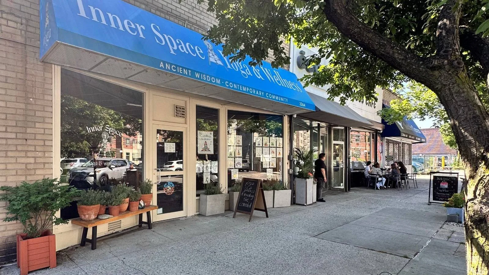 Inner Space Yoga & Wellness storefront on Johnson Avenue in Riverdale, Bronx, with a blue awning, workshop and class flyers in the windows, a chalkboard sign, and potted plants on a tree-lined sidewalk
