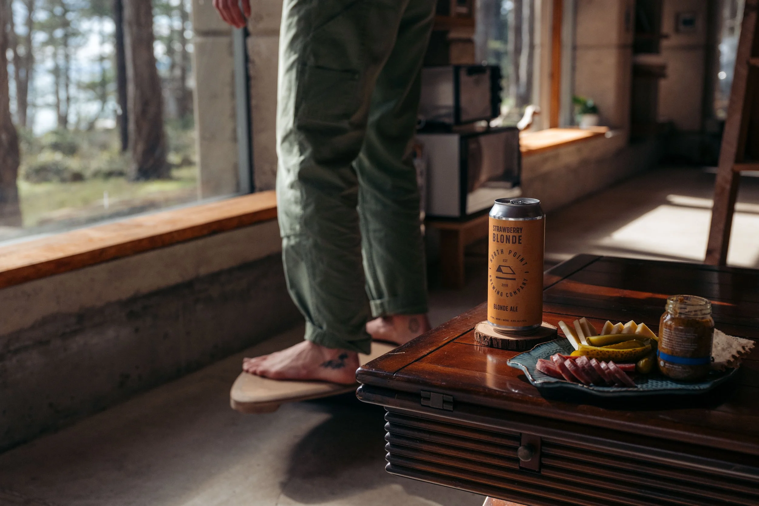 Indoor scene with a person standing on a skateboard near a large window, with a table holding a can of strawberry blonde ale, sliced vegetables, and a jar of mustard.