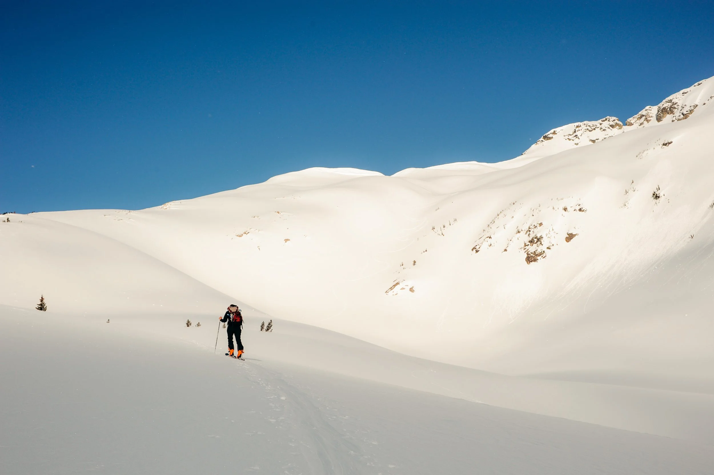 A person cross-country skiing through a snow-covered landscape with mountains in the background and a clear blue sky.