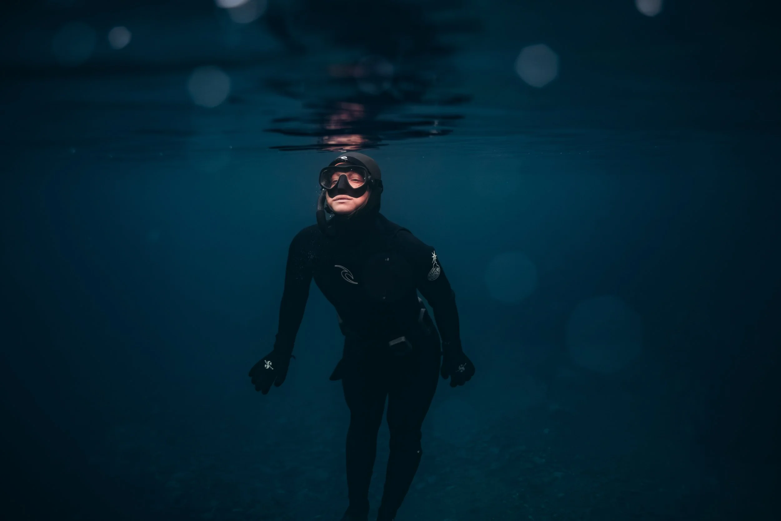 A person in black wetsuit, mask, and snorkel swimming underwater in the ocean.