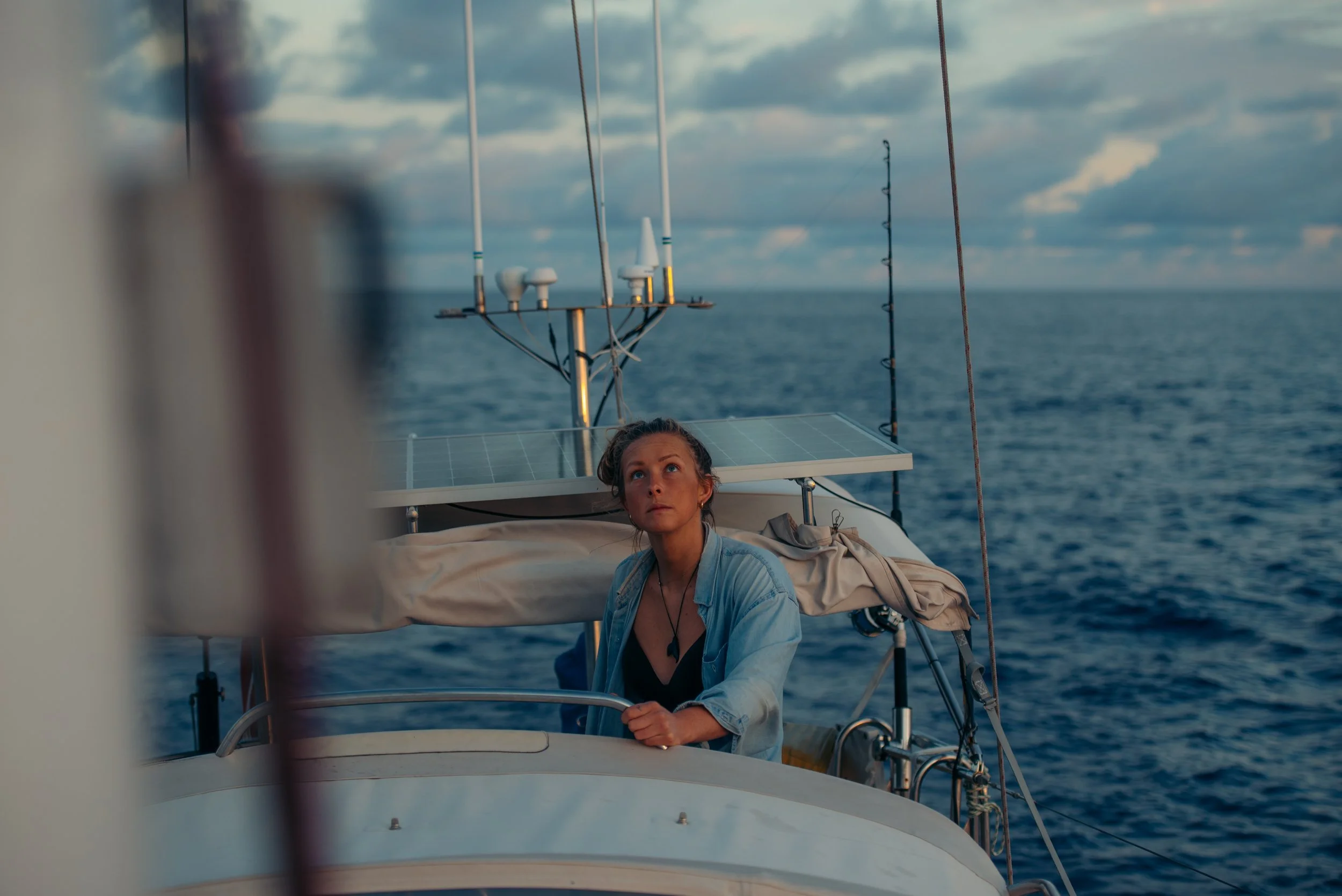 A woman on a boat at sea during sunset, looking up with a contemplative expression, with the ocean and cloudy sky in the background.