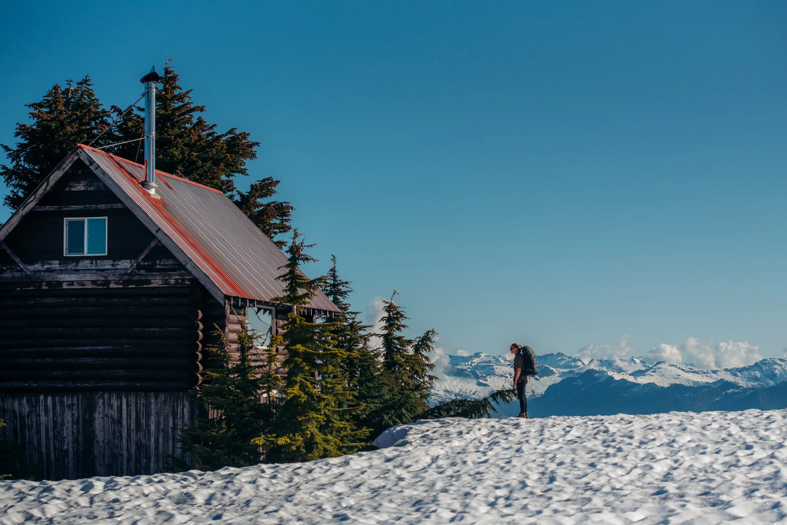 A person with a backpack standing on snow near a log cabin with pine trees and snow-capped mountains in the background.