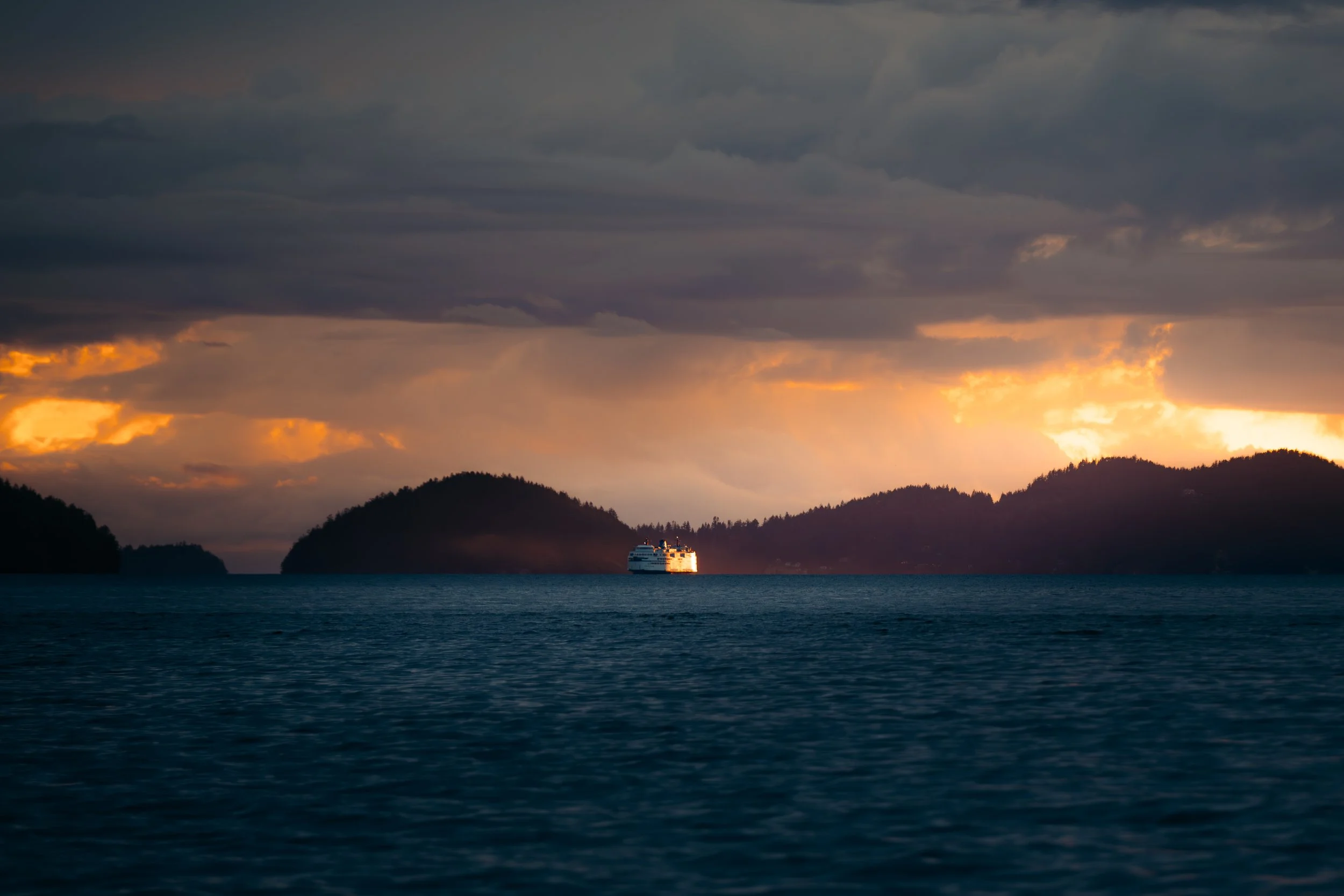 A large cruise ship sailing across a calm body of water with mountainous islands in the background during a dramatic sunset with dark clouds.