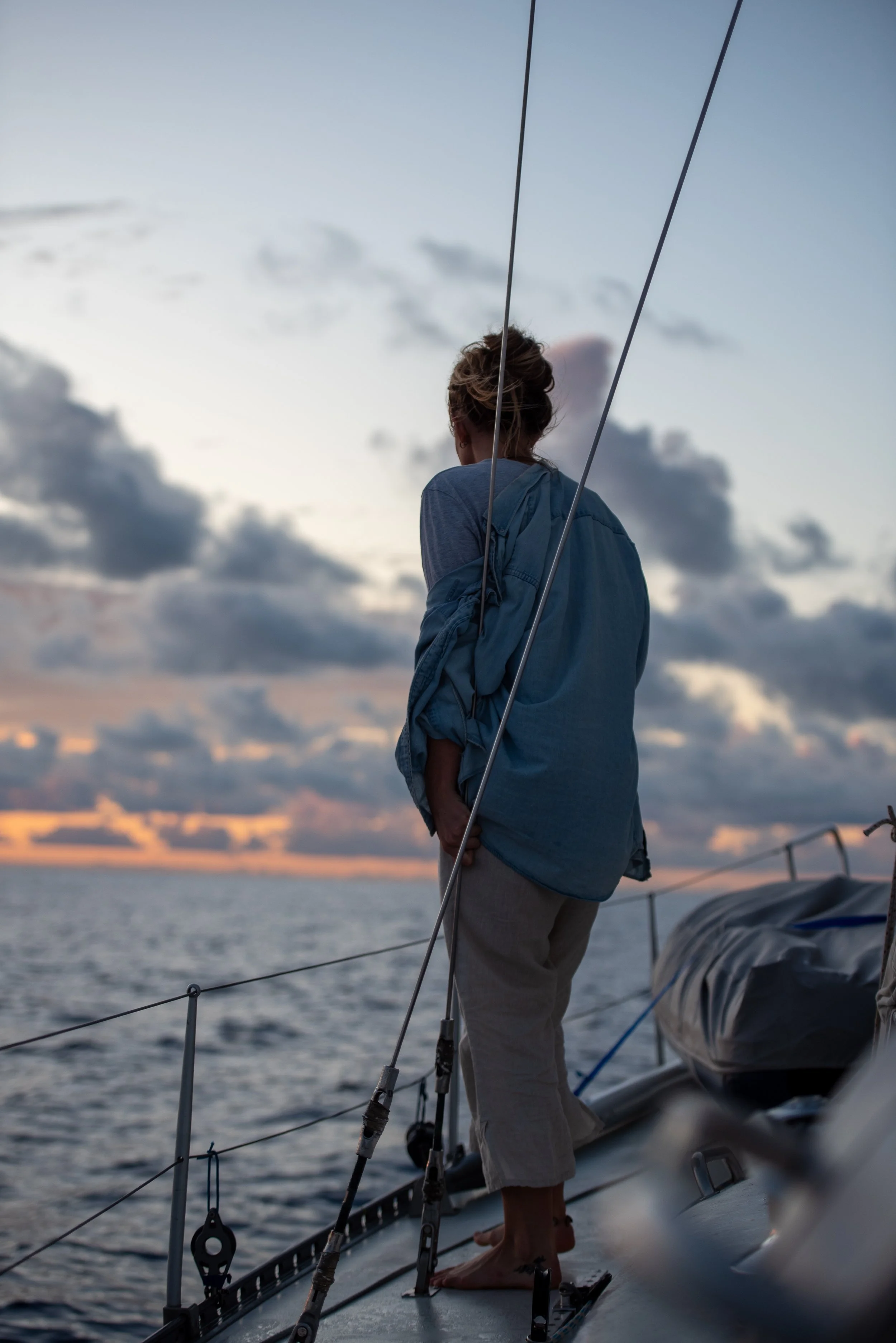 A woman standing barefoot on a boat deck at sunset, gazing at the ocean with clouds in the sky.