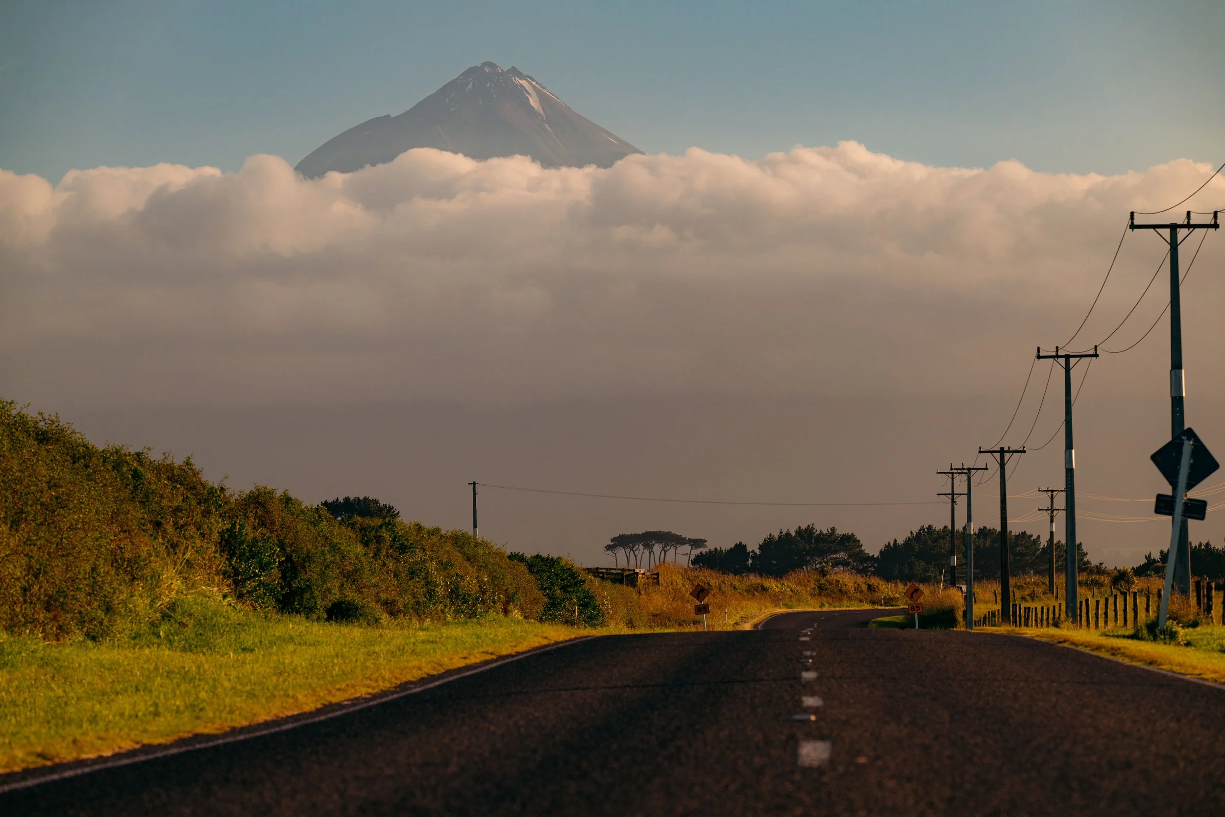 A winding country road with utility poles on the right, green bushes on the left, and Mount Fuji in the distance partially covered by clouds.