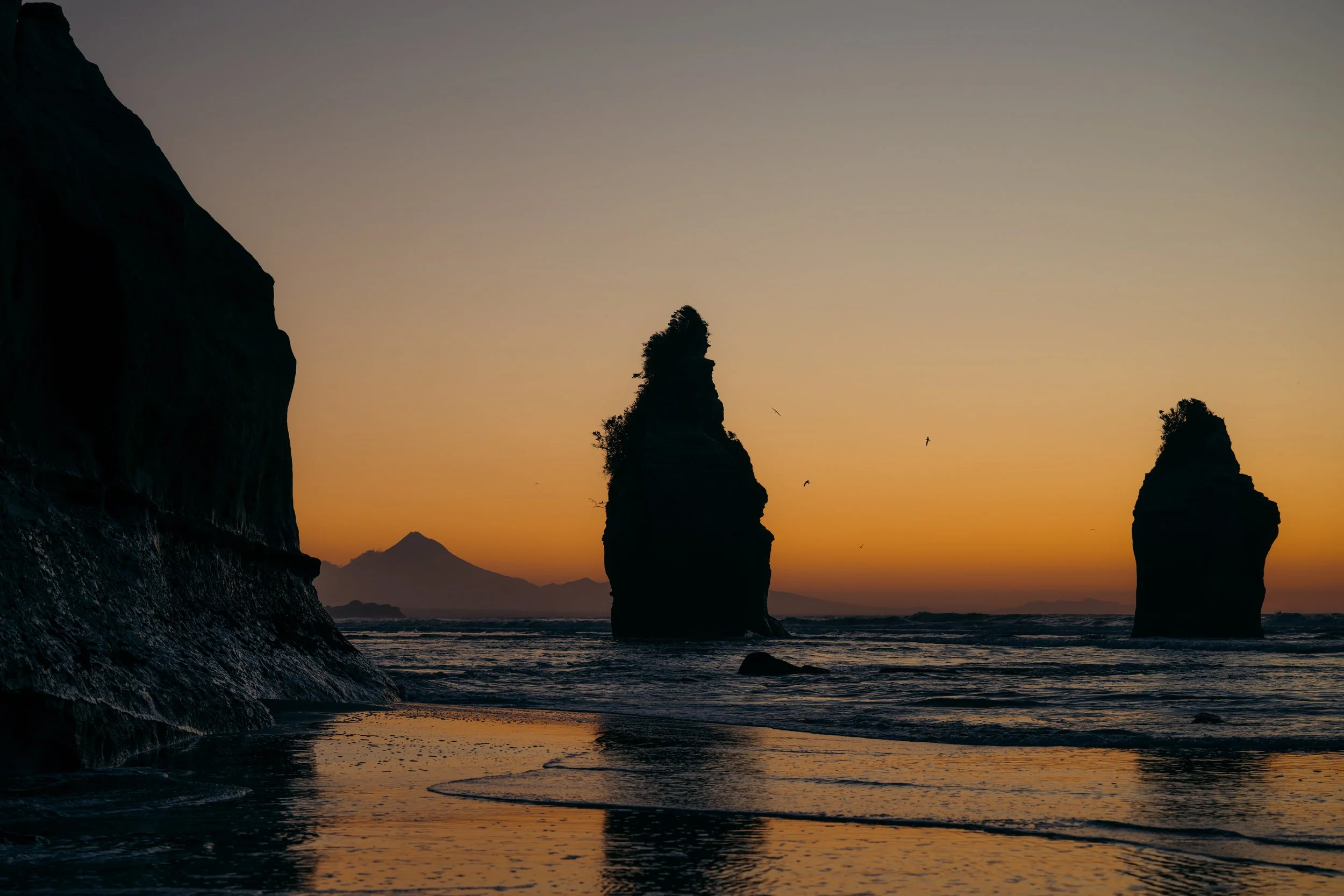 Silhouettes of large rock formations in the ocean at sunset, with a mountain in the background and seagulls flying in the sky.