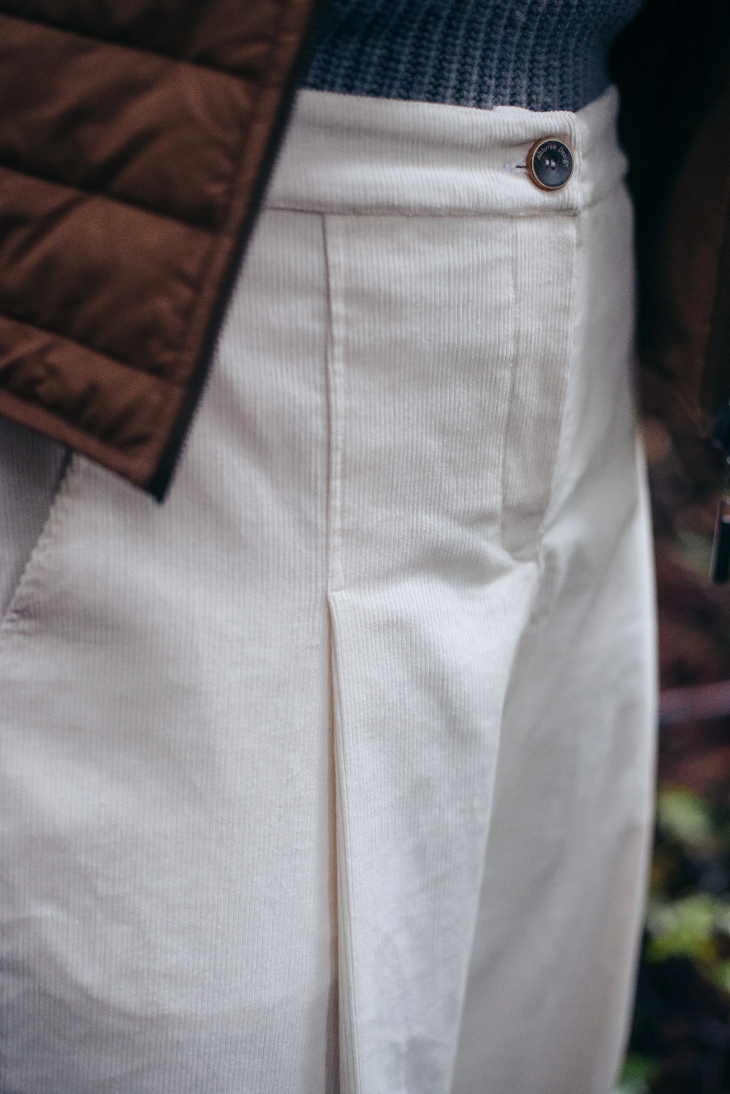 Close-up of a person wearing beige pants with a button close to the waistband, a blue sweater, and a brown quilted jacket.