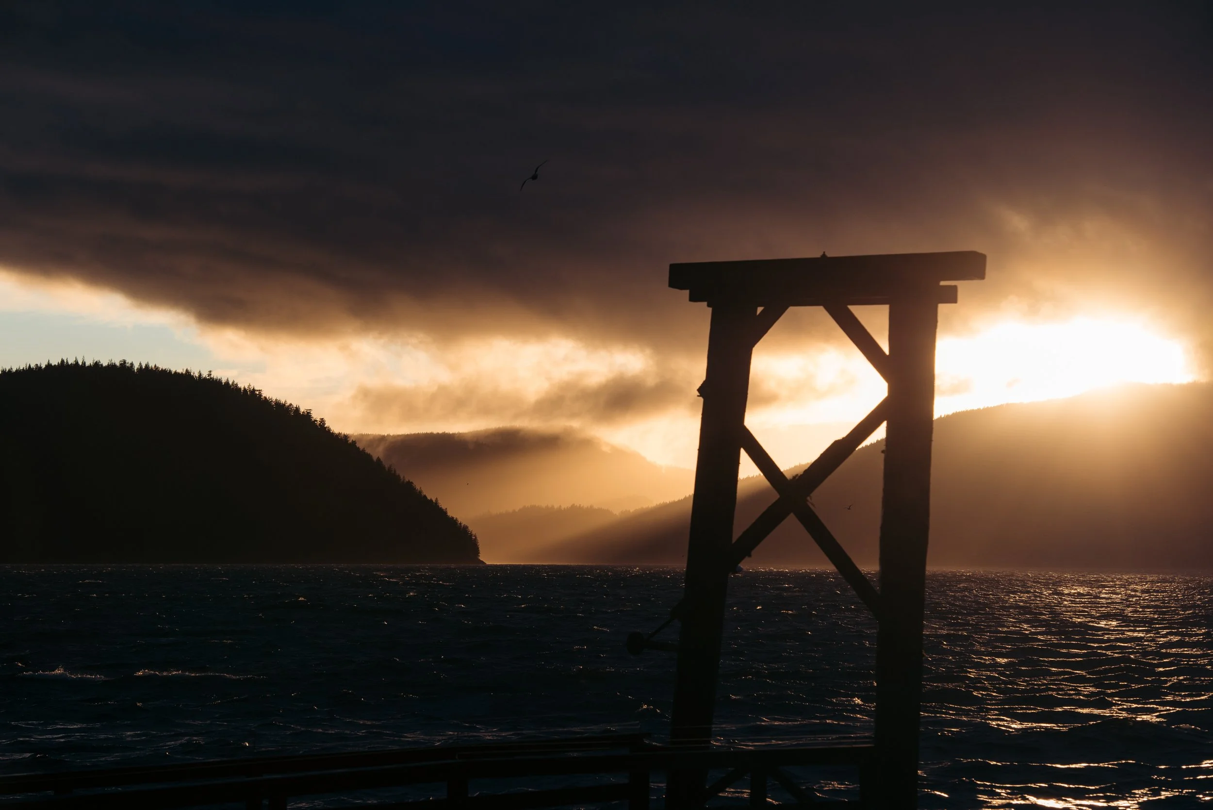 Silhouette of a wooden pier or structure beside a body of water with a mountainous landscape in the background and dark stormy clouds overhead, illuminated by the setting or rising sun.