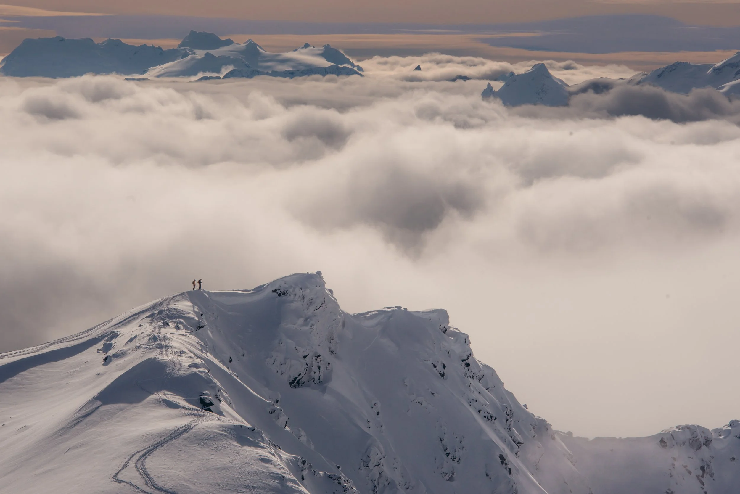 Two mountaineers standing on a snow-covered mountain peak above clouds and fog, with distant mountain ranges in the background.