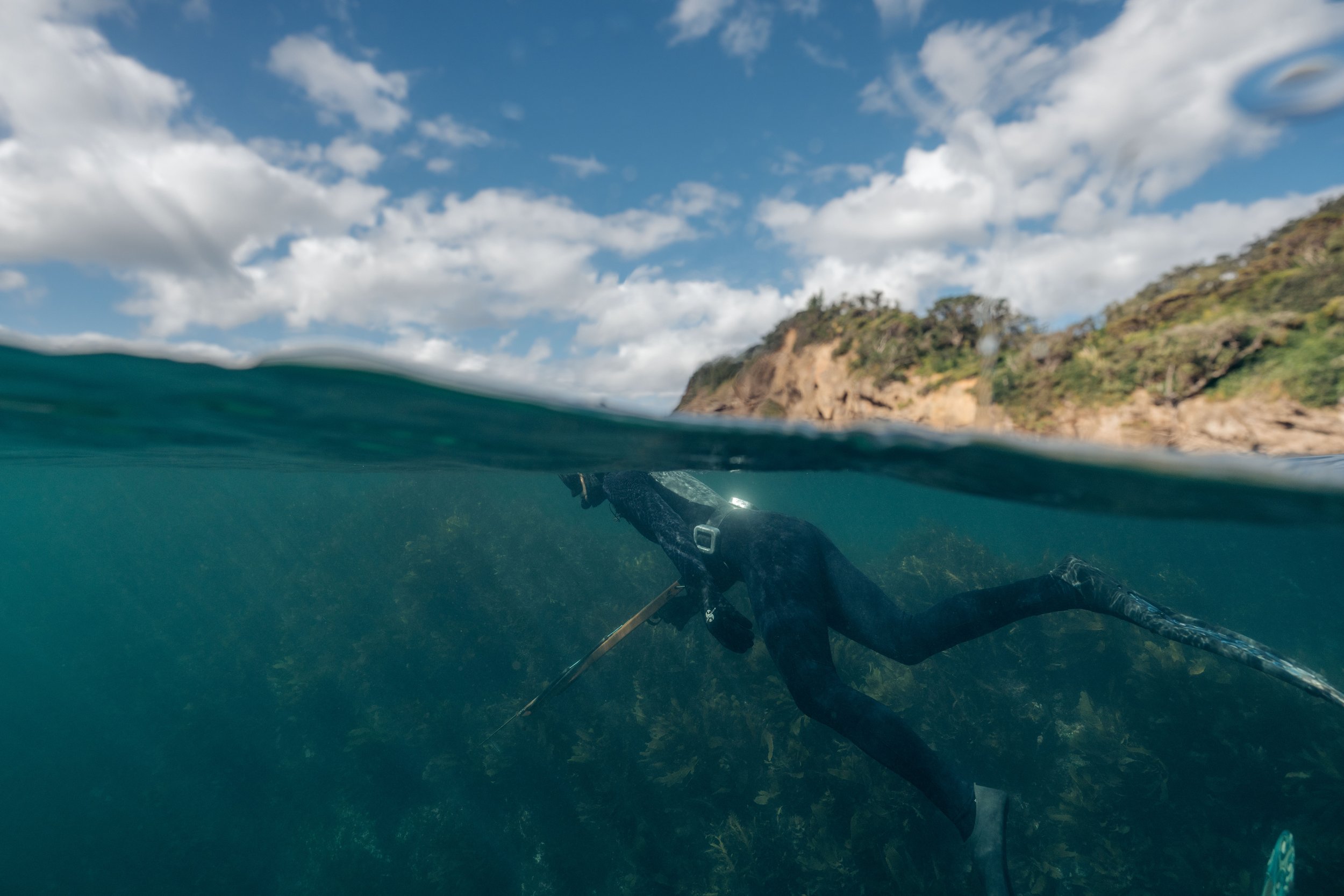 Half underwater, half above water view of a snorkeler swimming near a rocky shoreline with a blue sky and clouds above.