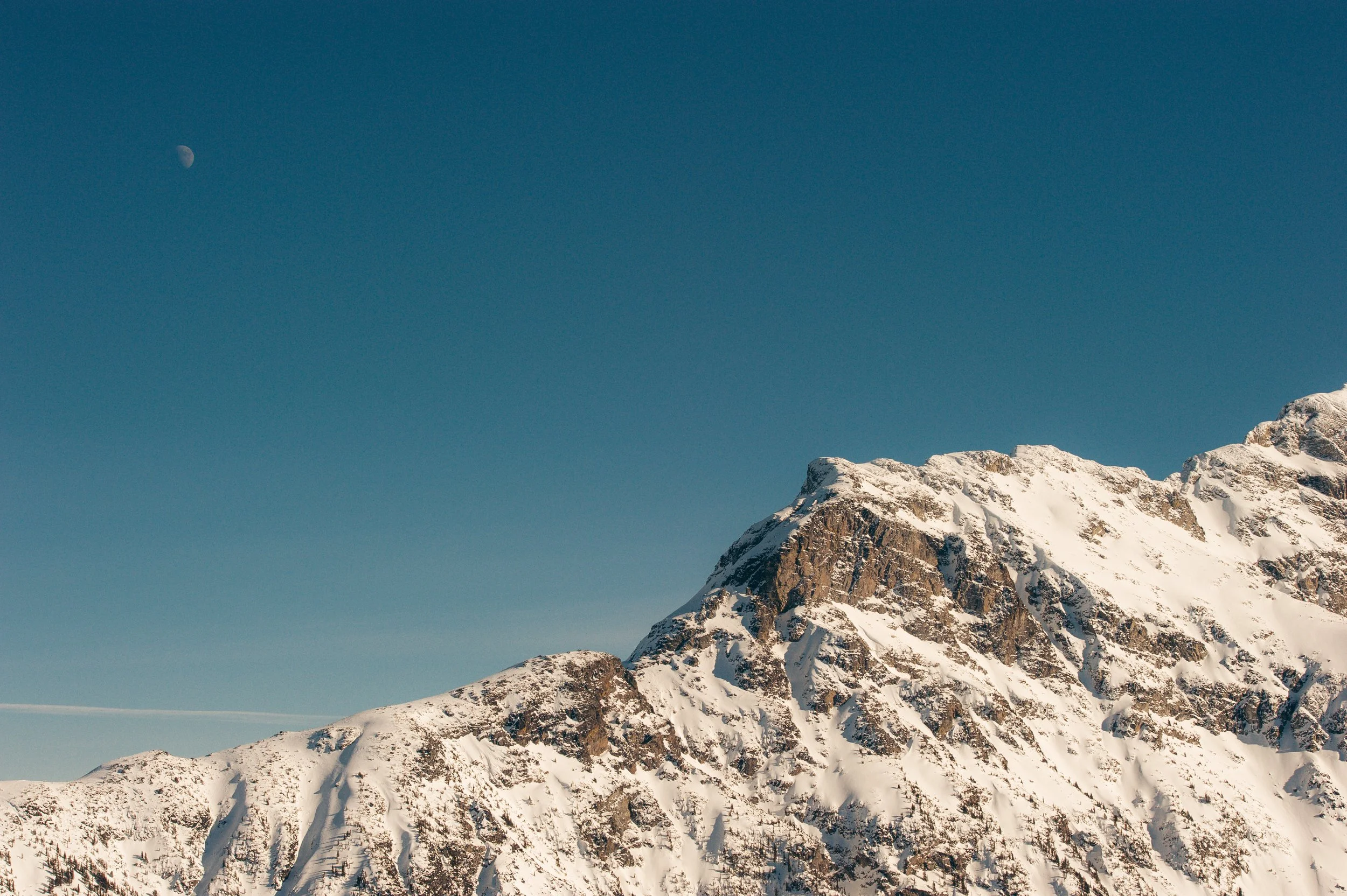 Snow-covered mountain range with blue sky and visible moon in the upper left corner.