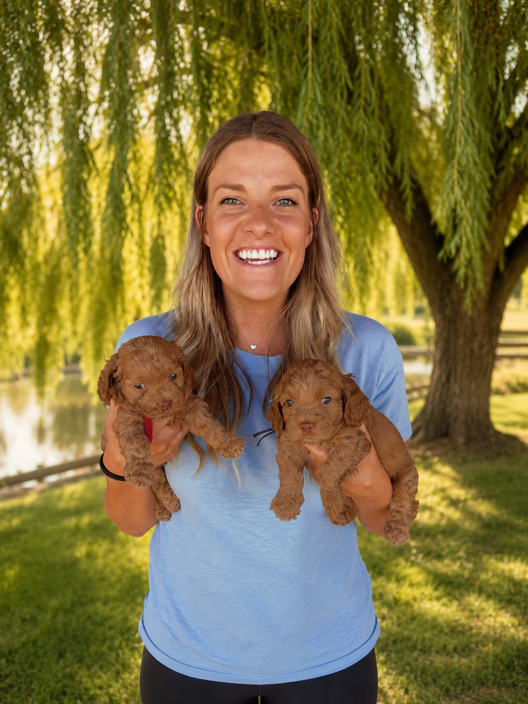 A smiling woman with long blonde hair holding a small fluffy puppy outdoors in a park with green trees and flowers.