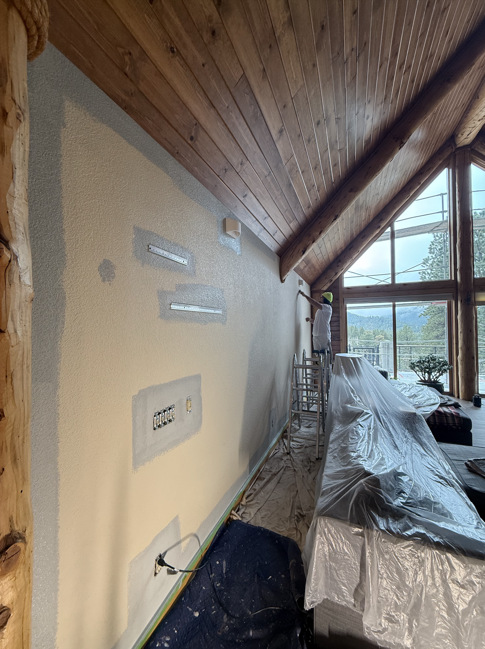 A worker is painting a wall in a room with a high, sloped wooden ceiling and large windows showing an outdoor landscape. Construction materials and covered furniture are visible.