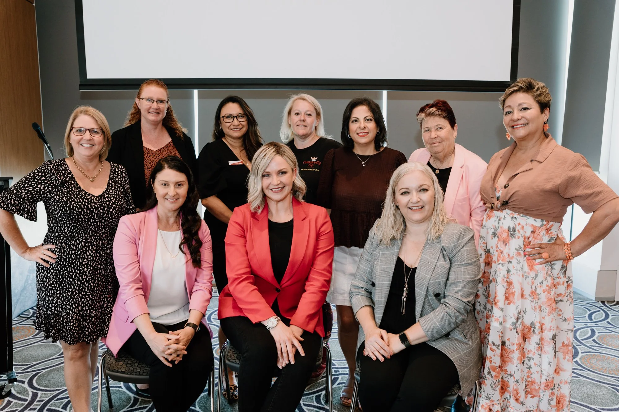 Group of eleven women posing for a photo in a conference room, some standing and some sitting, smiling at the camera, with a blank presentation screen behind them.