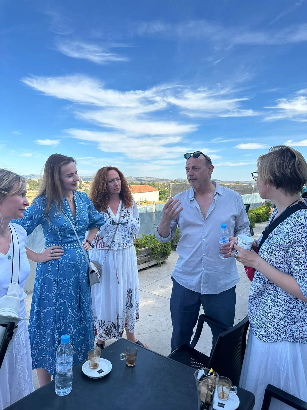 Group of five people having a conversation outdoors on a rooftop or terrace under a blue sky with clouds, with some drinking beverages and a table with drinks and water bottles in front of them.