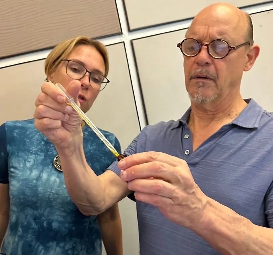 A man and woman, both wearing glasses, are involved in an activity where the woman is holding a dropper and applying a substance to a small object that the man is holding in his hand. They are focused on the task, possibly working on a craft or scientific experiment.