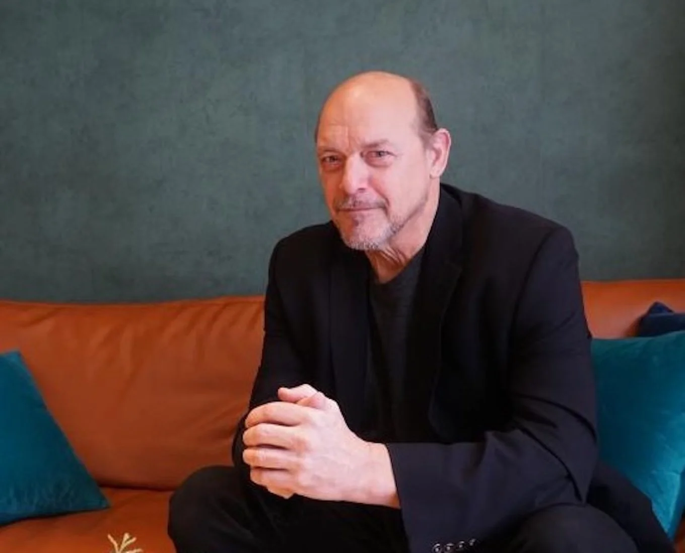 A man in a black blazer sitting on a brown couch with blue and teal pillows, hands clasped, in front of a dark green wall.