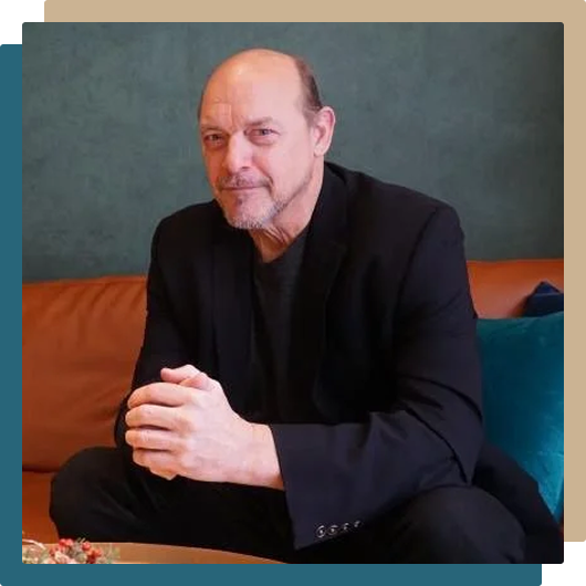 An older man with a beard and receding hairline sitting on a sofa with blue and brown cushions, wearing a black suit and black shirt, against a dark green wall.