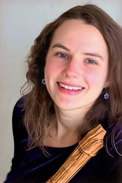 Close-up of a smiling young woman with wavy brown hair, wearing blue earrings and a dark top, holding a wooden object, against a plain light background.